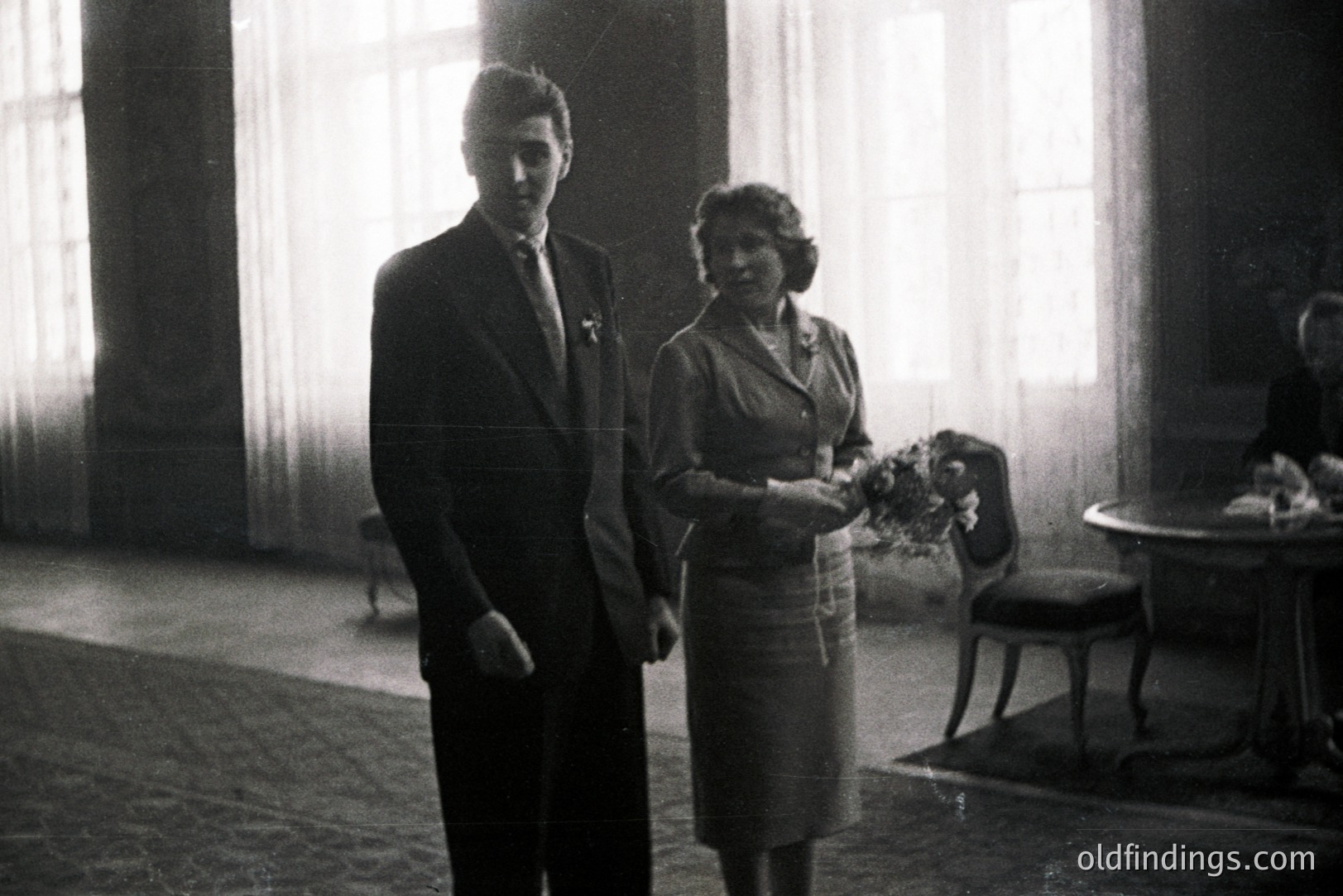 A formally attired couple stands near a window, likely at a wedding or celebratory event. The woman holds flowers; the man wears a suit & tie. Interior features include a round table & chairs. Appears to be 1950s-1960s photography.
