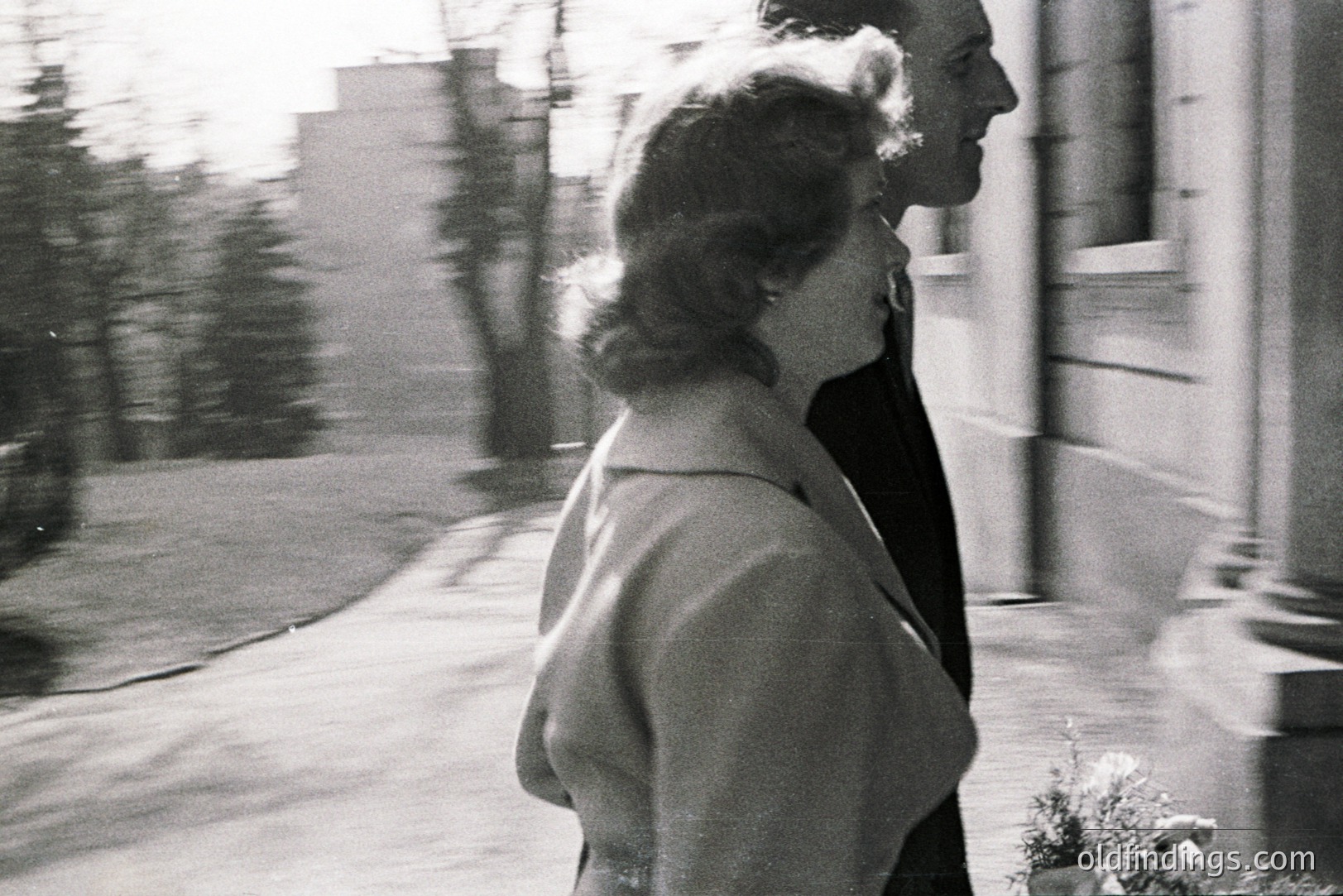 A formally dressed couple, possibly newlyweds, walk away from the camera along a paved path. Architectural details suggest a mid-century building facade. The blurred background hints at greenery and additional structures. Likely a candid moment captured in the 1960s. Potential stock photo reference for fashion or event design.