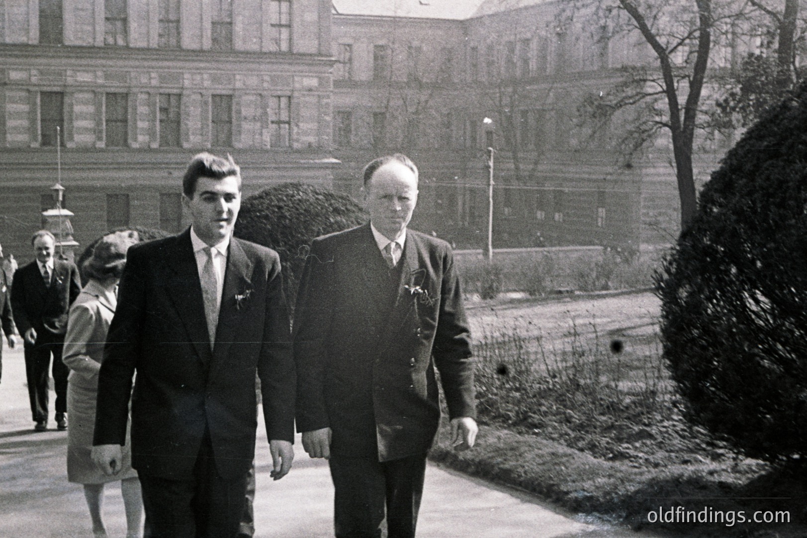 Two formally attired men walk along a paved path lined with manicured shrubs, a grand building visible in the blurred background. The photograph captures a moment of understated elegance, likely mid-20th century. The subjects appear to be engaged in conversation, a sense of formality and perhaps importance conveyed through dress and posture.