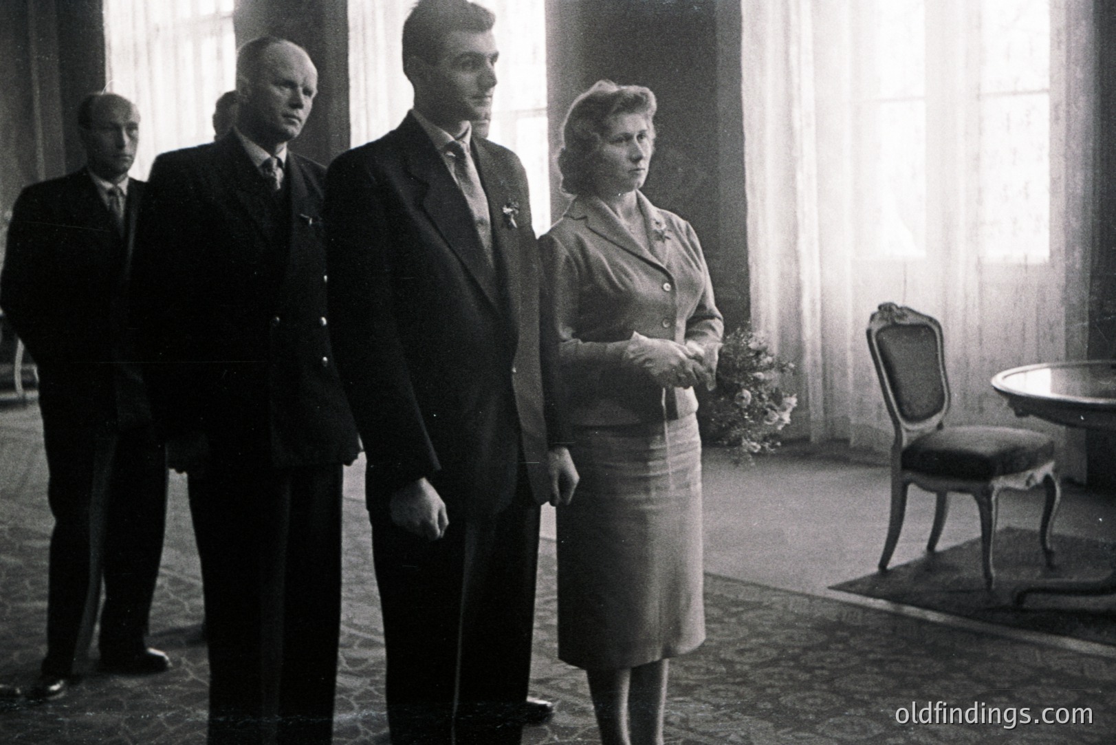 Formal portrait shows three men and one woman in dark suits and a matching skirt set, likely a diplomatic or military gathering. Interior features ornate patterned carpeting, a draped window, and dark wood furniture. Appears to be from the 1940s or 1950s.