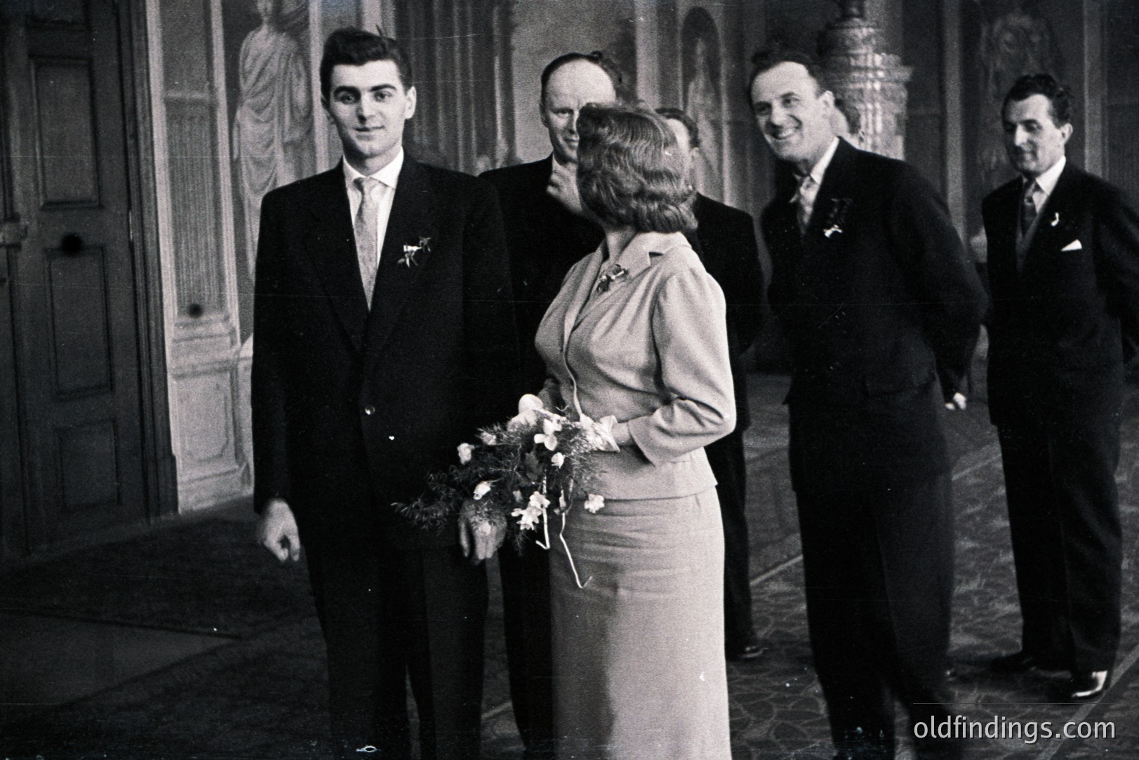 A young bride in a pale suit carries a bouquet, posed with four formally dressed men, likely groomsmen. The setting appears to be a grand hall, with a glimpse of a statue visible behind. Formal attire and hairstyles suggest a 1960s or 1970s wedding. Architectural details indicate a European location, possibly a stately home or castle. Likely stock photo reference for vintage weddings.