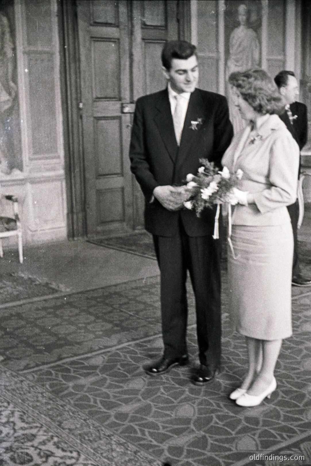 A formally dressed couple stands facing the camera in a grand interior. The groom, in a dark suit, holds flowers; the bride wears a matching suit and carries a bouquet. A large decorative urn is visible behind them, and a partially visible figure stands to the right. Elegant flooring & architecture suggest a 1960s European wedding.