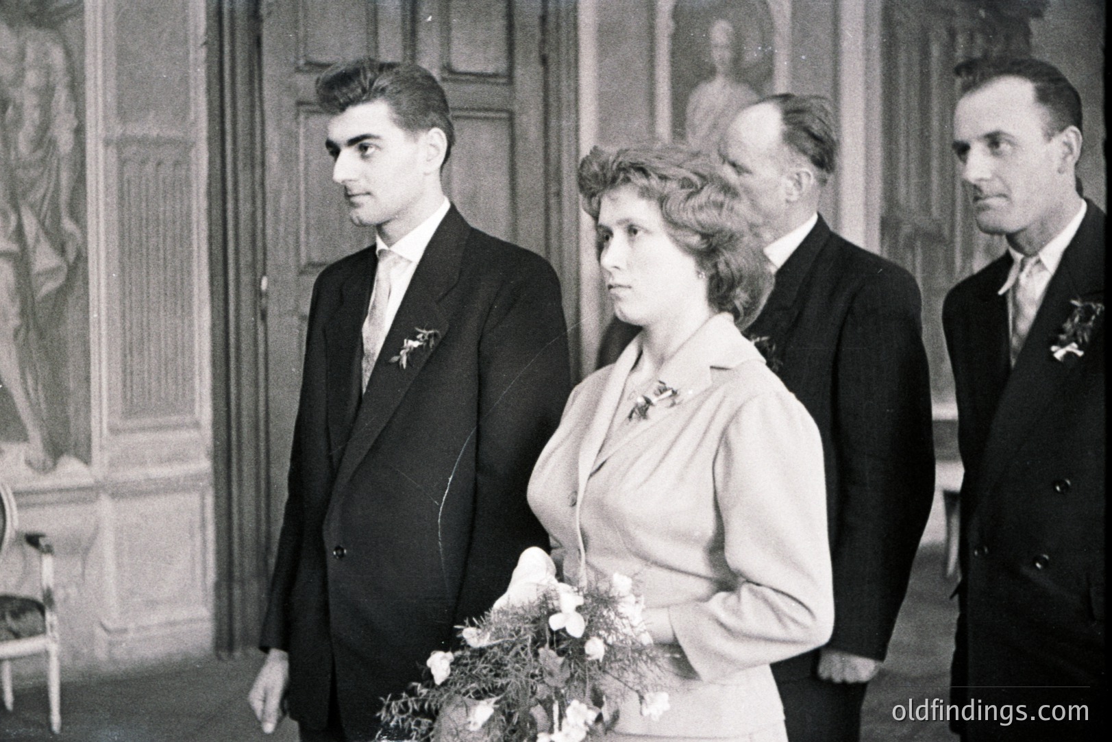 Formal portrait showcases a wedding party. A young couple stands at center, with two older men flanking them. The bride holds a bouquet. Architectural details suggest a stately interior, possibly a manor or estate. Likely 1950s, potentially Eastern European.
