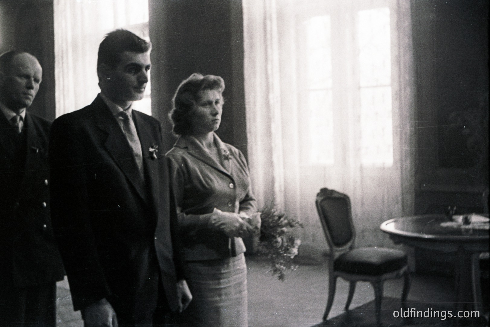 Formal portrait of a couple and a gentleman in a room with large windows. The man wears a suit and tie, she a suit with gloves, both appearing solemn. Interior suggests an official or ceremonial setting, possibly a wedding. Likely 1950s-1960s. Architectural details are minimal.