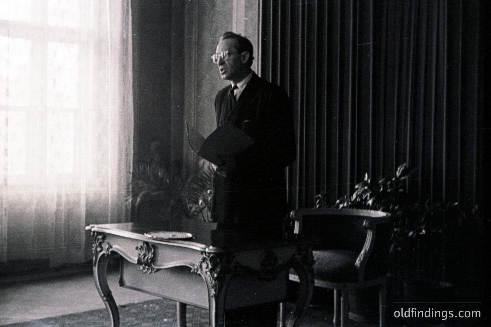 A man in a suit and glasses stands holding papers near an ornate desk, likely in an office. Strong directional light illuminates the scene. Interior features heavy drapes and a potted plant. Suggests a formal workplace setting, possibly 1950s-1970s. Black and white photography.