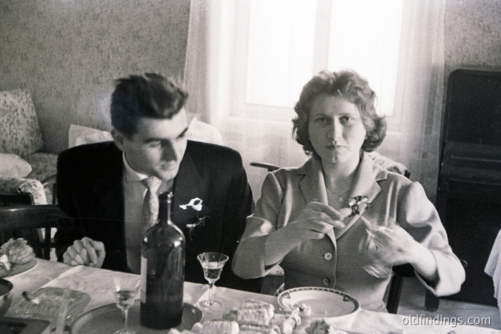A formal, black-and-white portrait captures a couple seated at a table laden with food and drink. The man, in a dark suit, gazes forward; the woman, in a tailored jacket, appears to be examining a small object. Likely a domestic interior, 1960s. May be valuable for design and historical context.