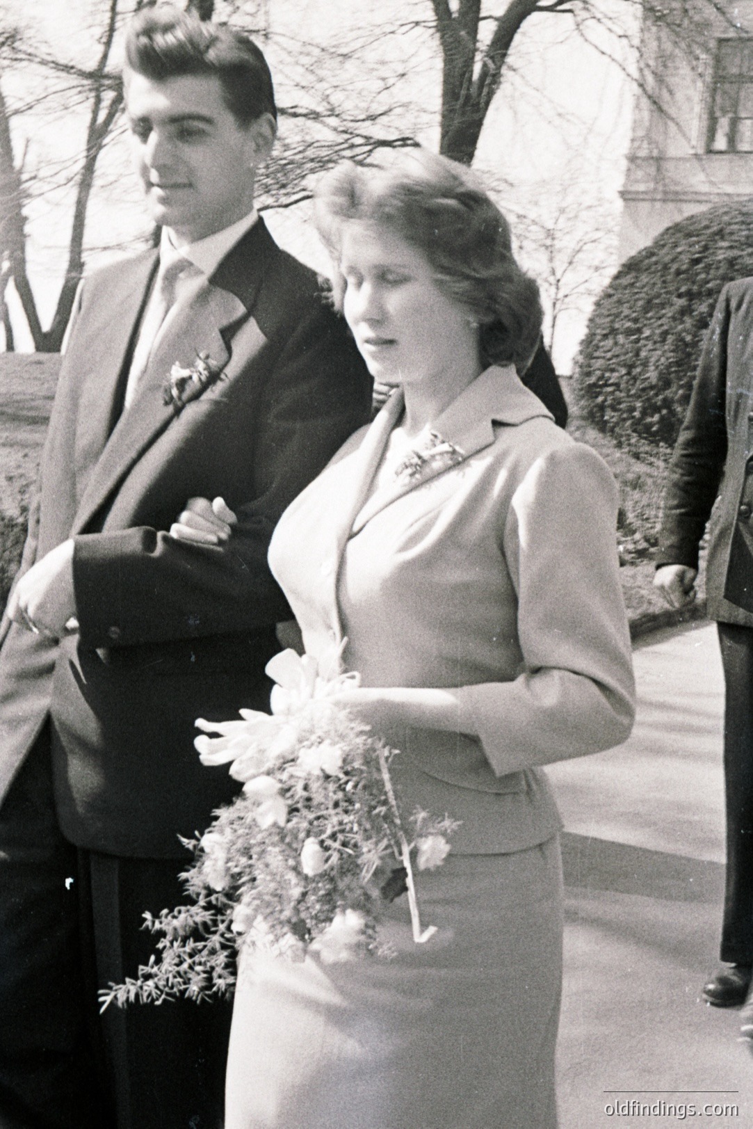 A posed black and white portrait captures a formally dressed couple, likely during a wedding. The woman holds a bouquet of flowers; the man stands beside her in a dark suit and tie. Architectural details suggest a stately building in the background. Circa 1960s-1970s.