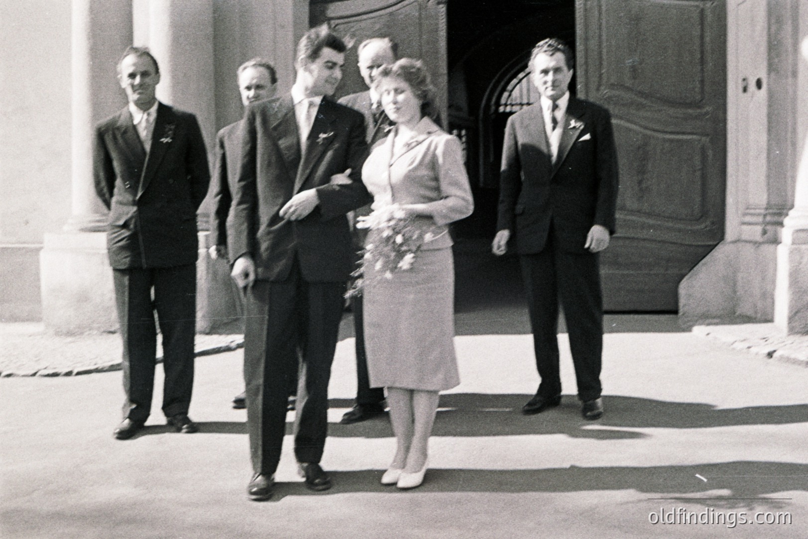 Formal portrait of a wedding party outside a grand building. Bride in a knee-length dress and floral bouquet stands between four formally dressed men in suits and ties. Architectural details suggest a European setting, possibly 1950s-1970s. Classic, potentially valuable for design or historical research.