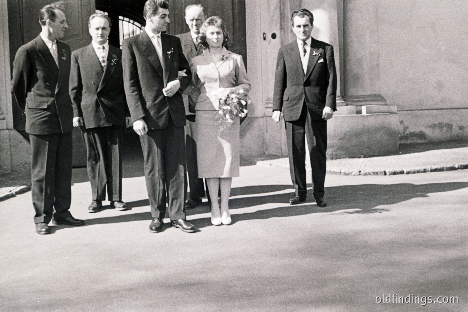 Black & white photo depicts a wedding party exiting a building. Five figures: groom, two groomsmen, the bride, and another man (likely a father or official). Formal attire, mid-length dress. Likely 1950s-1960s. Architectural background suggests a European location. Possible stock photo reference for vintage weddings.