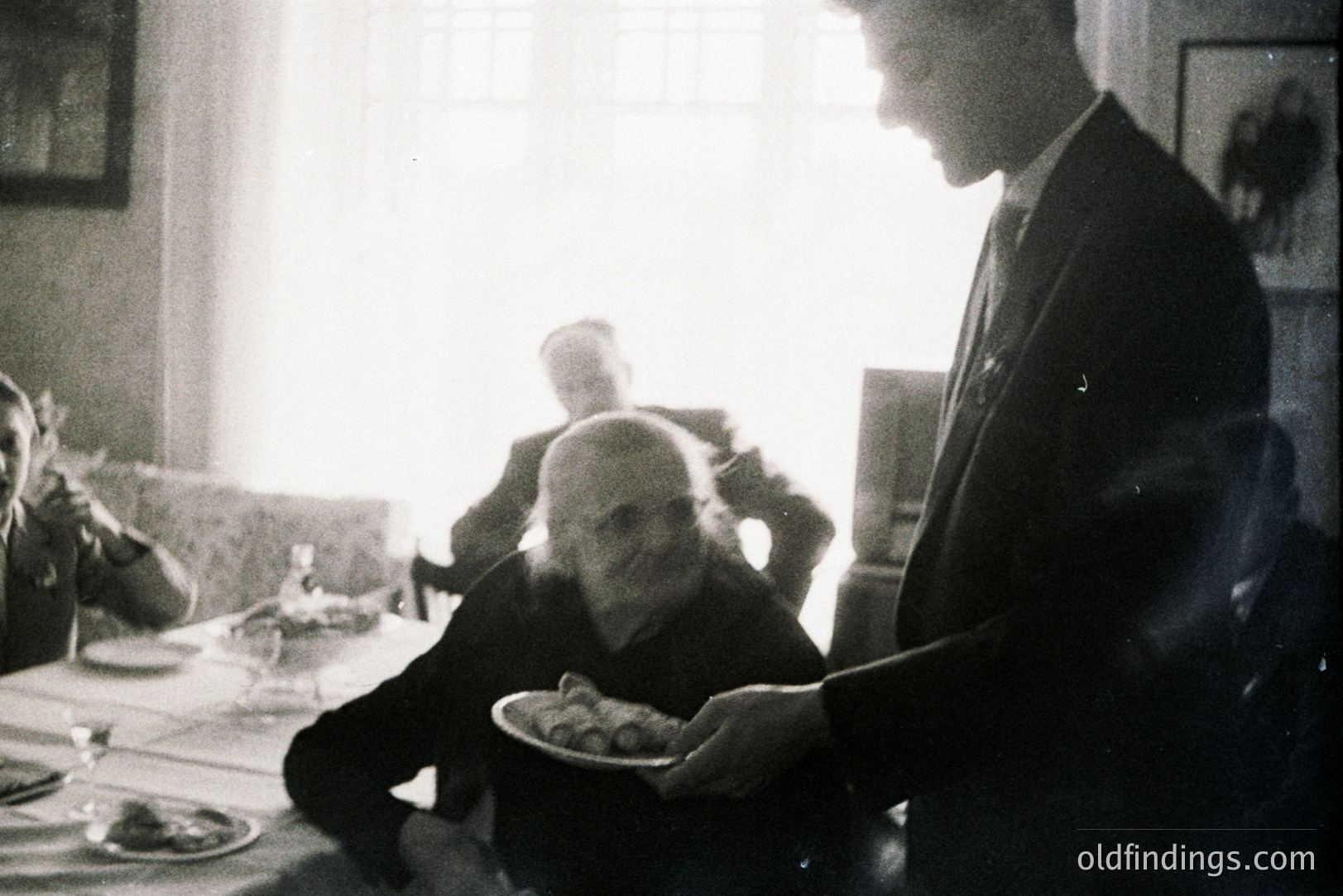 B&W image depicting a dimly-lit interior scene. A server in a suit presents a plate of food to a seated, elderly gentleman with a bald head. Two other figures are partially visible in the background. Suggests a formal gathering or domestic setting. Likely mid-20th century.
