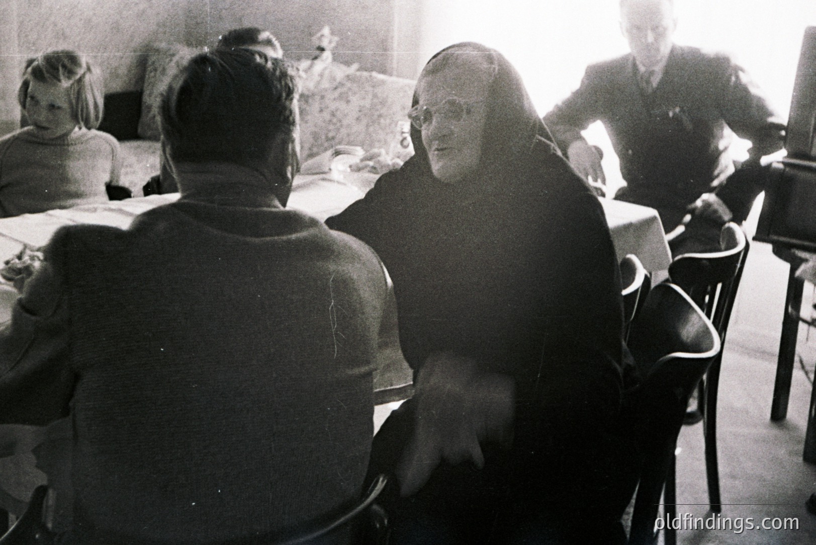 A candid, intimate moment: three figures sit around a table covered with a white cloth. A nun, wearing a dark habit and glasses, appears to be speaking to a man whose back is to the camera. A young girl is visible in the blurred background. Likely taken in Europe, 1950s-1970s. Potential stock value for historical or religious contexts.