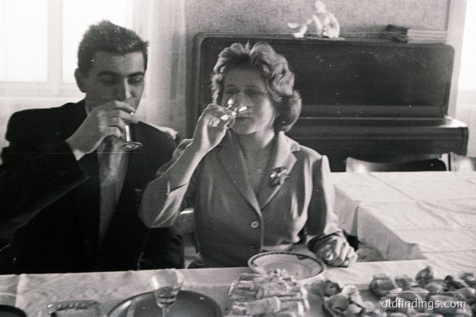 A formally dressed couple shares a moment at a table laden with food and wine glasses, likely a celebratory dinner. The woman holds a wine glass, the man drinks. Appears to be a domestic interior with a decorative mantelpiece and still life arrangement.