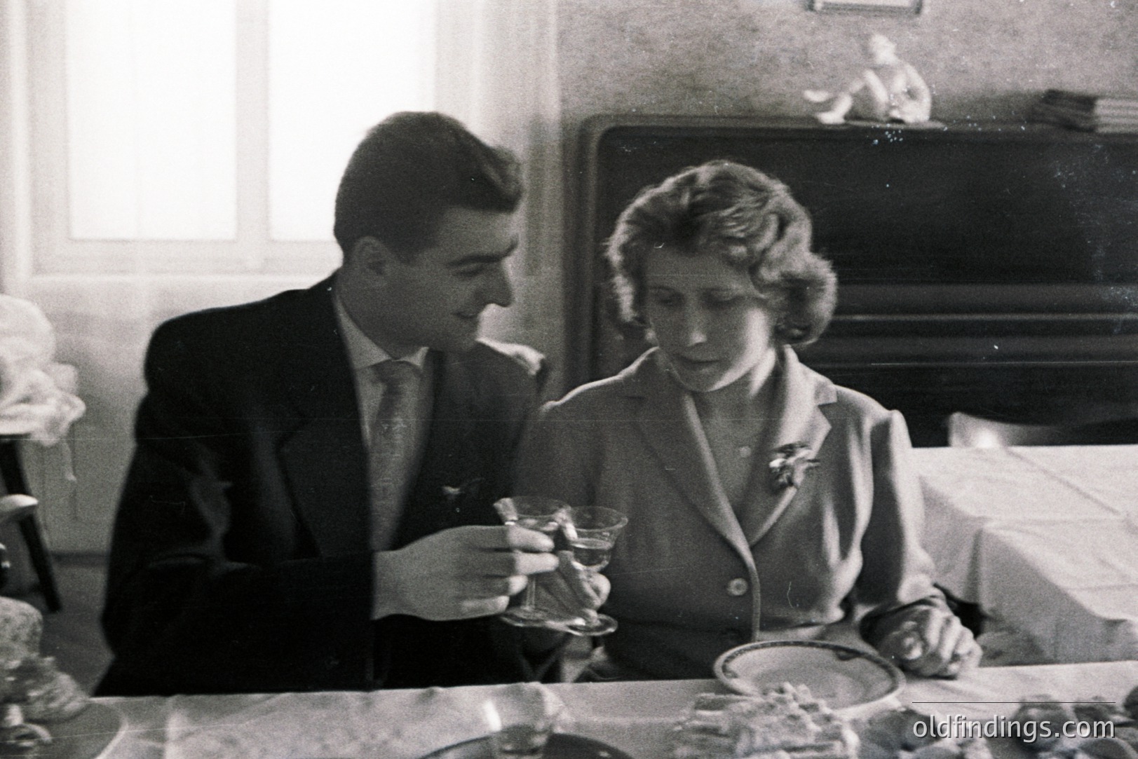 A formally dressed couple shares a toast in a domestic interior, likely a dining room. The man raises two glasses; the woman gazes downwards. A plate of food sits on the table covered with a white tablecloth. Fireplace and framed artwork visible in background.