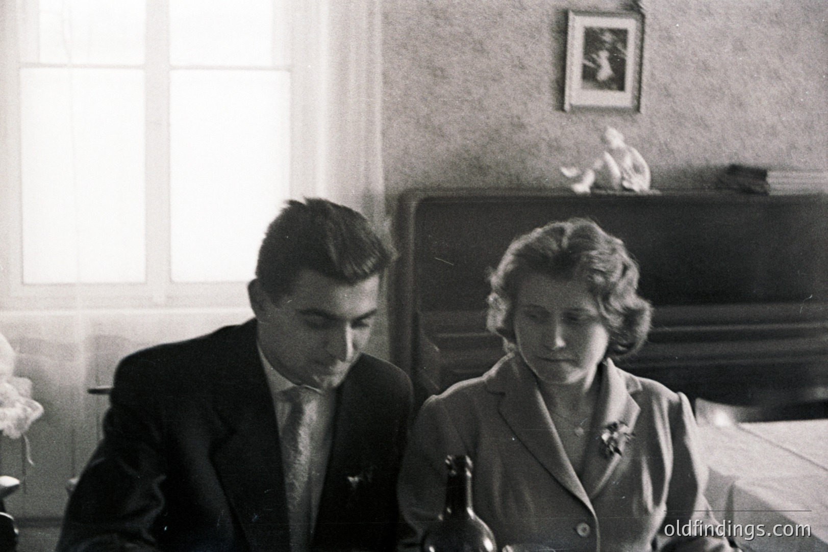 A formally dressed couple sits at a table covered with a white cloth. The man in a suit and tie appears contemplative; the woman wears a tailored dress with a floral brooch. A bottle sits on the table between them. Interior scene, likely 1950s or 60s.