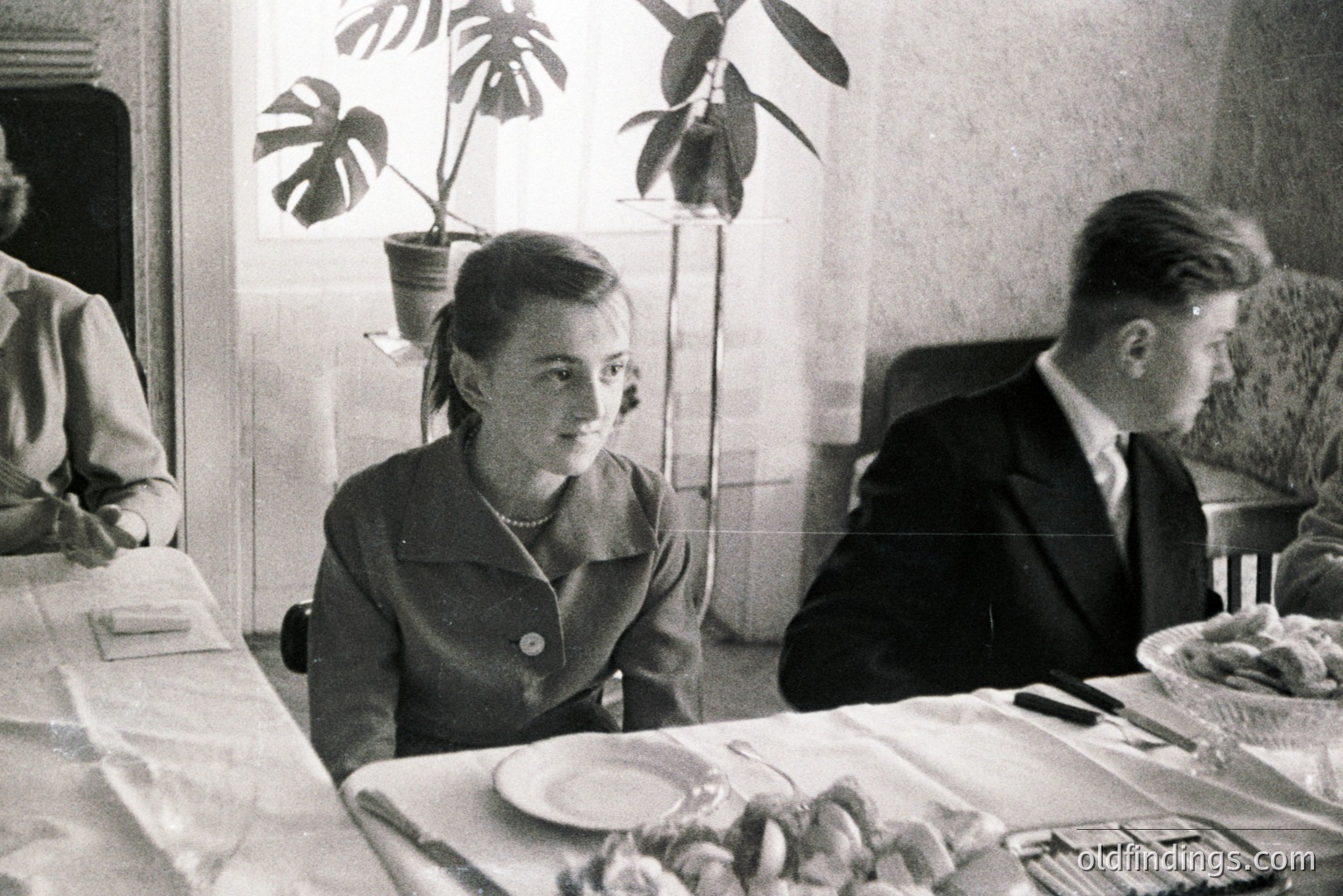 A seated woman with a short, styled haircut gazes forward during what appears to be a formal dinner or gathering. A young man sits beside her, also formally dressed. A table laden with food dominates the foreground. Indoor plants are visible in the background, suggesting a well-appointed home. Likely 1950s, potentially European.
