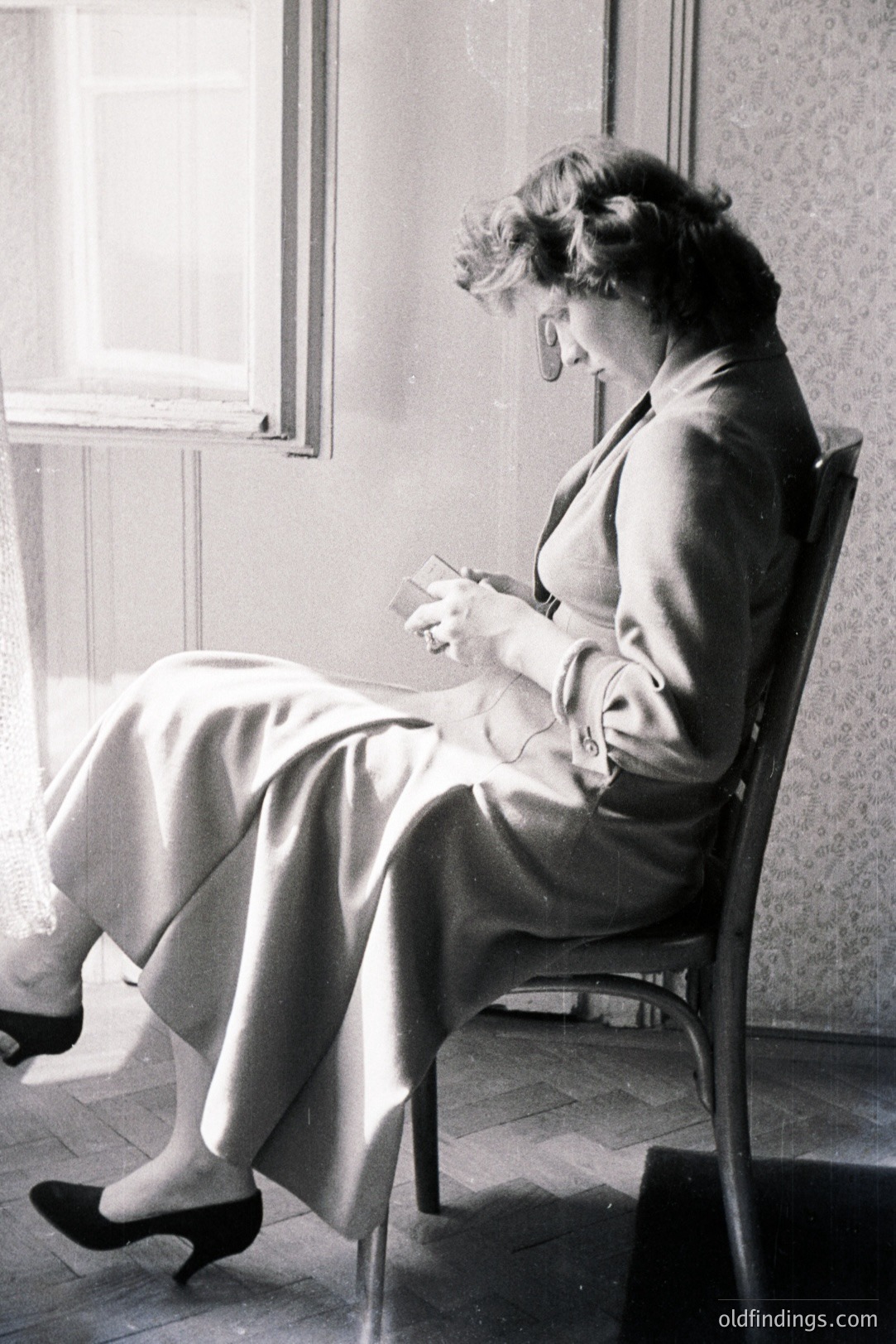 A woman seated in a chair, engrossed in reading a book. She wears a wide-legged suit and dark heels. Natural light illuminates the scene, highlighting the wood-paneled walls and the subject’s focused expression. Likely dating from the 1960s or 70s. A study in quiet contemplation.