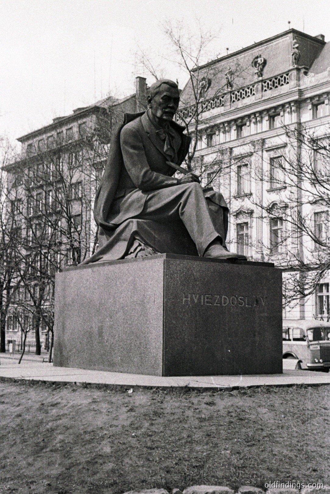 A black and white image depicts a bronze statue of a seated man in a suit and cloak atop a stone pedestal. The inscription "HVIEZDOSLAV" is visible on the base. A grand, ornate building with multiple windows stands in the background. Likely a memorial or public art installation.