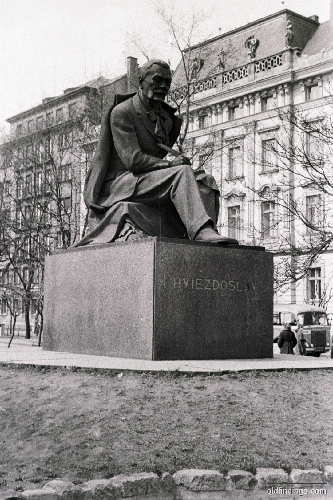 Seated bronze statue of a man in a suit, appearing contemplative, rests on a dark stone plinth inscribed "HVIEZDOSLAV." A large, ornate building and vintage car are visible in the background. Likely a public monument in a European city, 1950s-1970s.