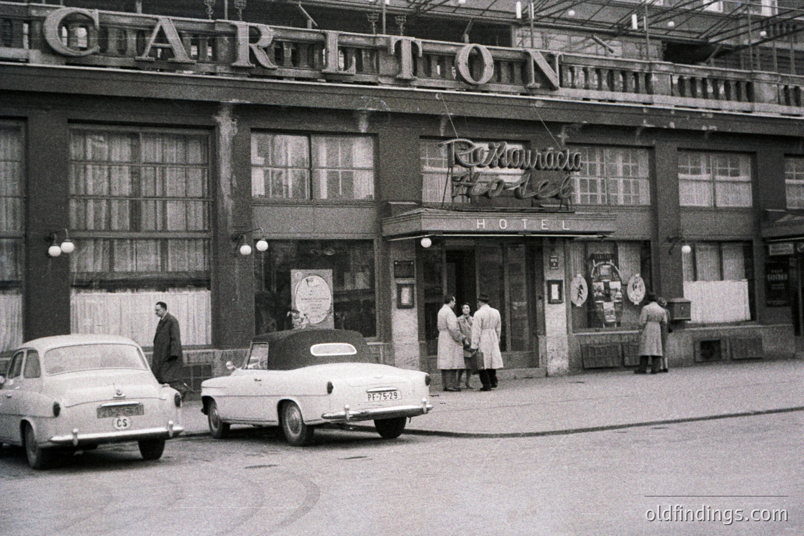 Hotel entrance, likely 1960s, features a prominently signed facade and classic cars parked along the street. Three figures are arriving, suggesting guest activity. Art Deco architectural elements are visible. Location appears to be Eastern Europe.