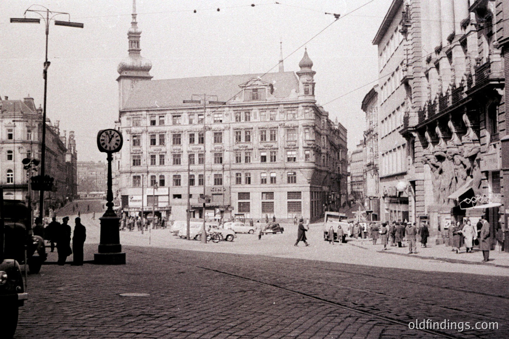 Historic street scene. Ornate, multi-story building with a central clock tower dominates the composition. Cobblestone street with vintage automobiles and pedestrians. Likely a European city, circa 1950s-1970s, judging by the cars and dress. Detailed architecture and urban planning.