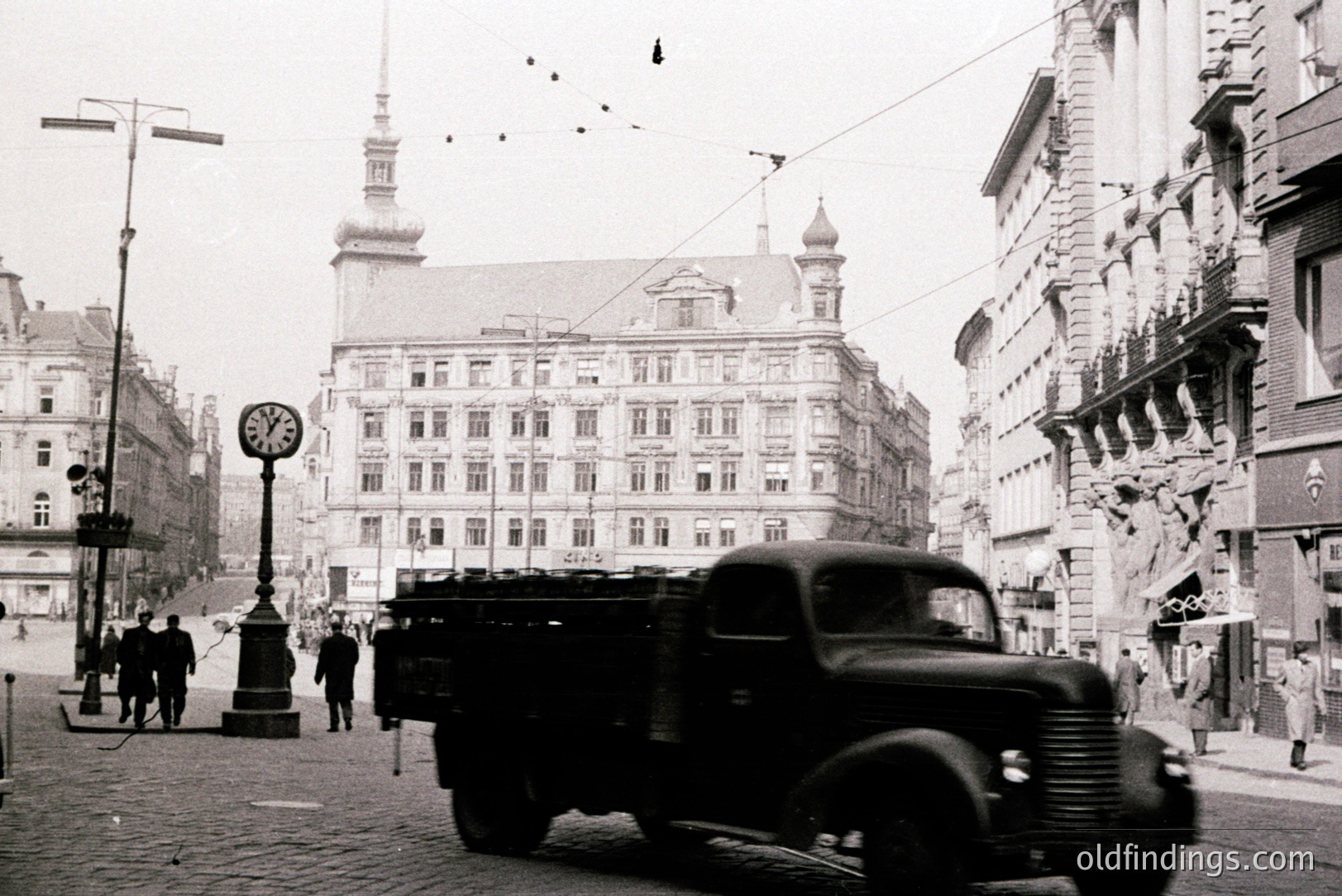 A bustling street scene, likely post-war Europe, features a military transport truck dominating the foreground. Ornate, multi-story buildings line the paved road, punctuated by a clock tower. Several figures are visible on the sidewalk, creating a snapshot of daily life amidst a backdrop of architectural detail.