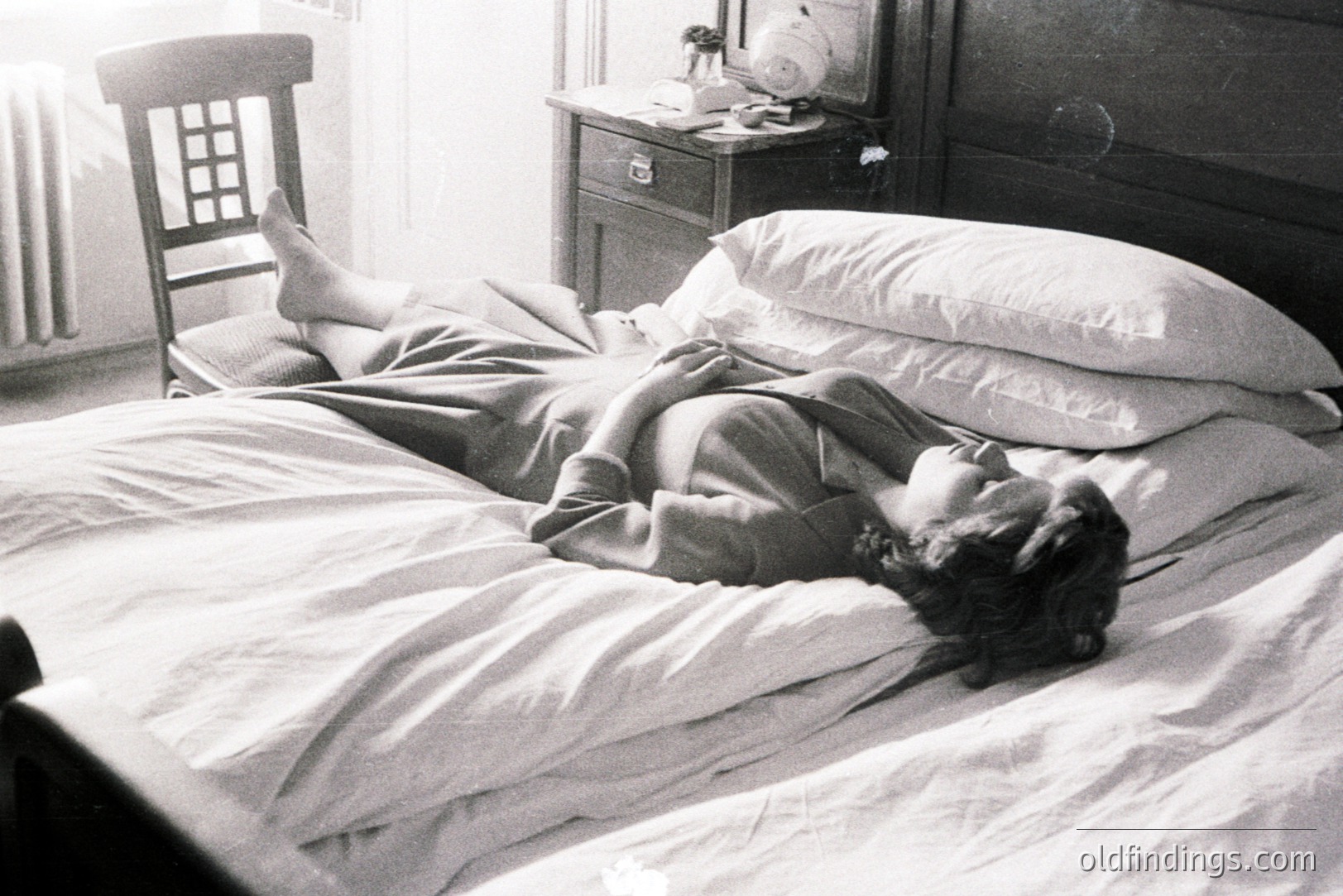 A young woman reclines on a heavily draped bed, appearing relaxed or asleep. A wooden chair sits nearby, and a bedside table holds a lamp and vase. Likely a 1960s lifestyle or fashion photograph, showcasing domesticity. The image's composition evokes quietude and privacy.
