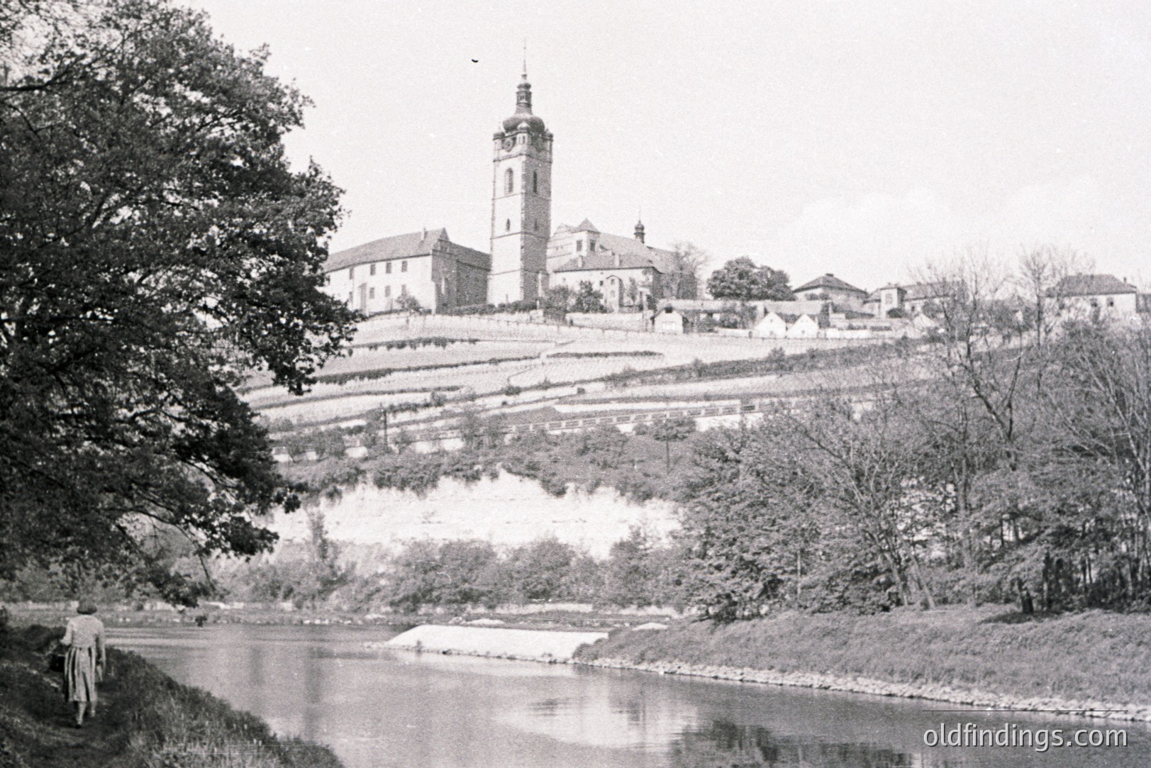 A historic townscape with a prominent church tower dominates this vintage image. Terraced slopes rise toward the buildings, hinting at agricultural use. The river in foreground reflects the scene. Likely from the early 20th century, possibly 1900s-1930s.