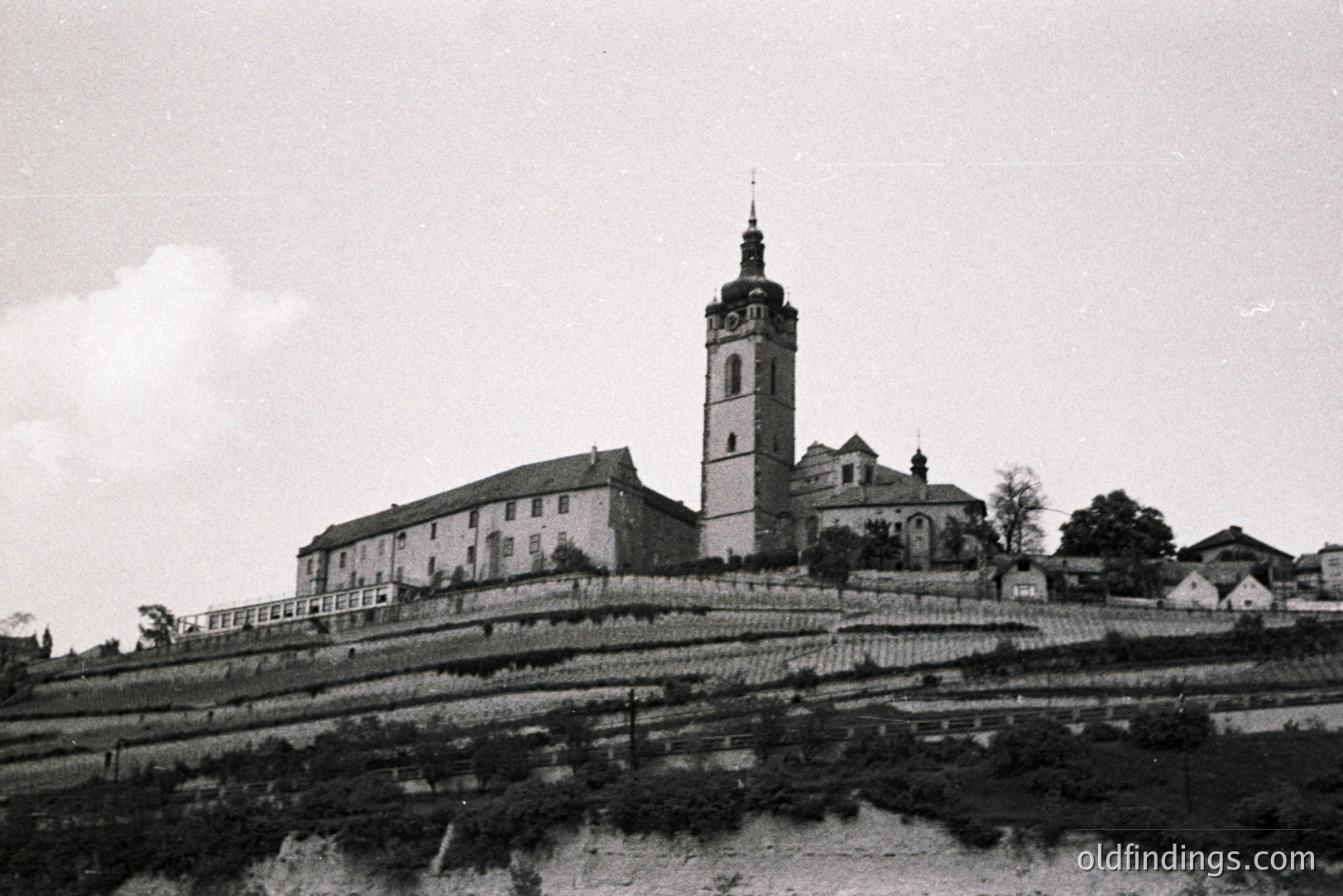A hillside terraced landscape leads up to a large stone monastery with a prominent clock tower. The building displays multiple windows and a complex roofline. Likely a European location, possibly Central Europe. Appears to be an architectural study or tourist view.