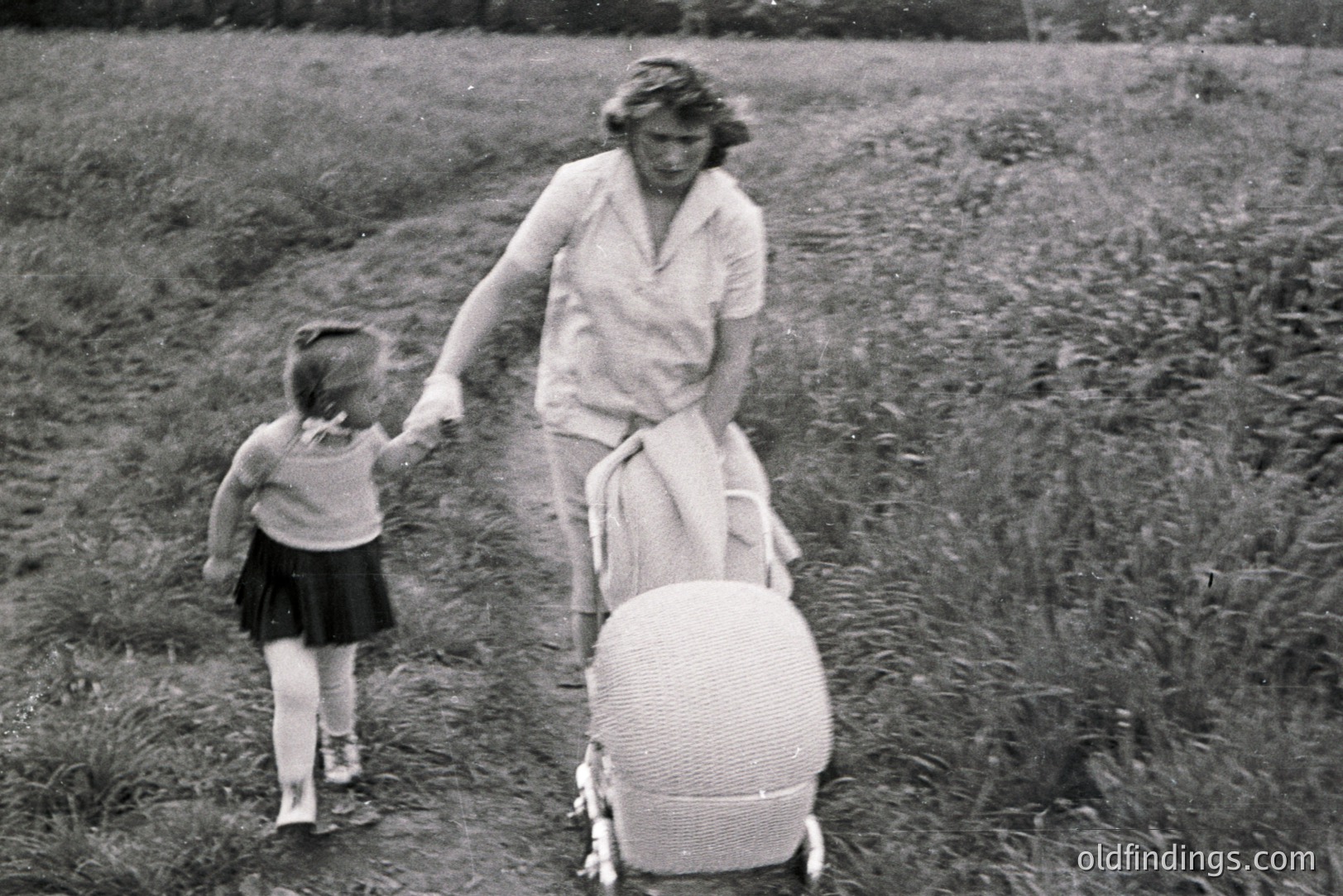 Black & white image: a mother & young girl walk along a path bordered by tall grass. The mother wears a light-colored dress, while the girl sports a pleated skirt & short sleeves. A woven stroller accompanies them. Likely a family outing, c. 1950s-1960s. A nostalgic, domestic scene.