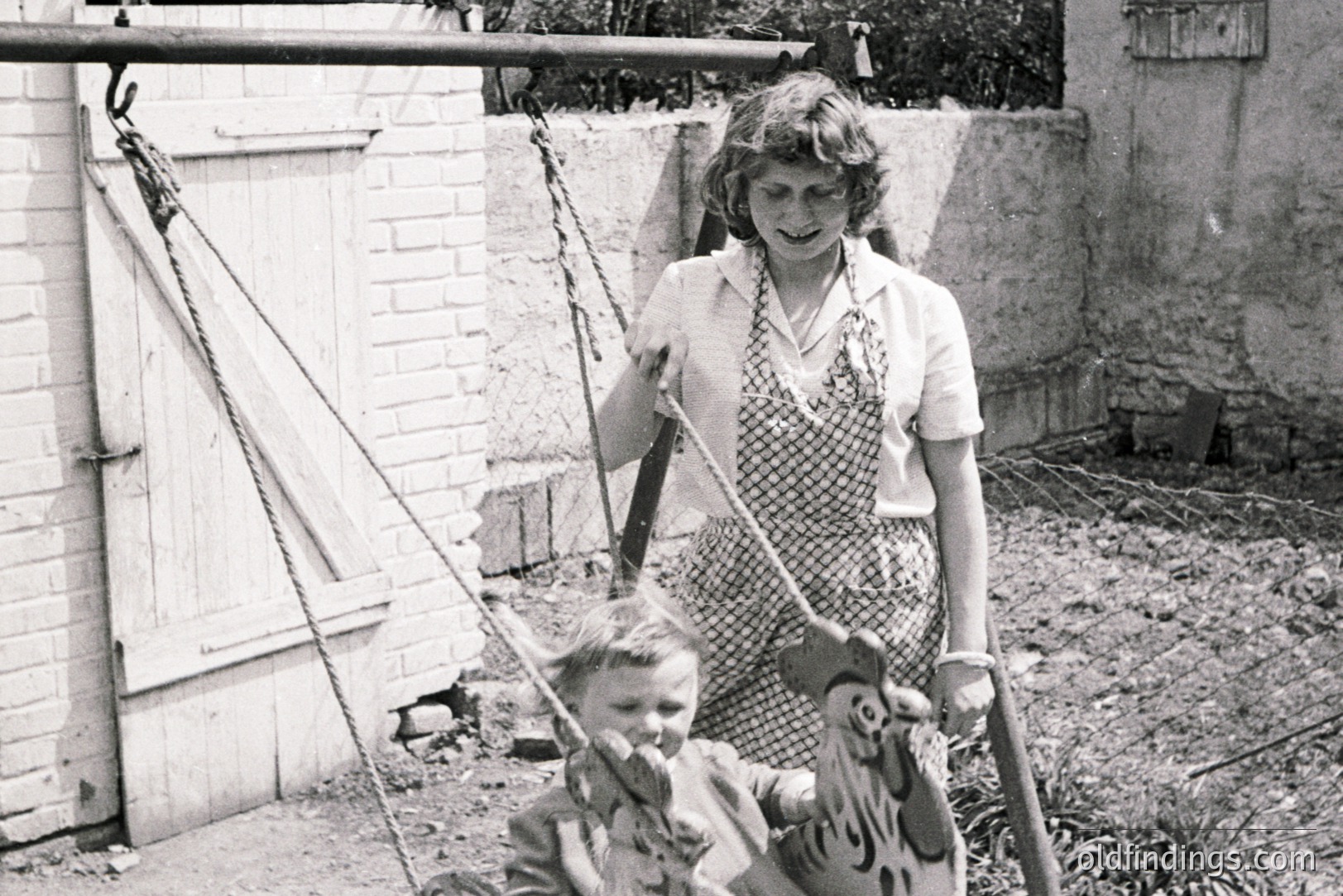 A woman watches a young boy on a whimsical, painted wooden tiger swing. Mid-century backyard scene shows a brick wall and overgrown vegetation. Likely a family snapshot, circa 1950s, offering a glimpse of domestic life and childhood play. Could be valuable for design and nostalgia-themed stock.