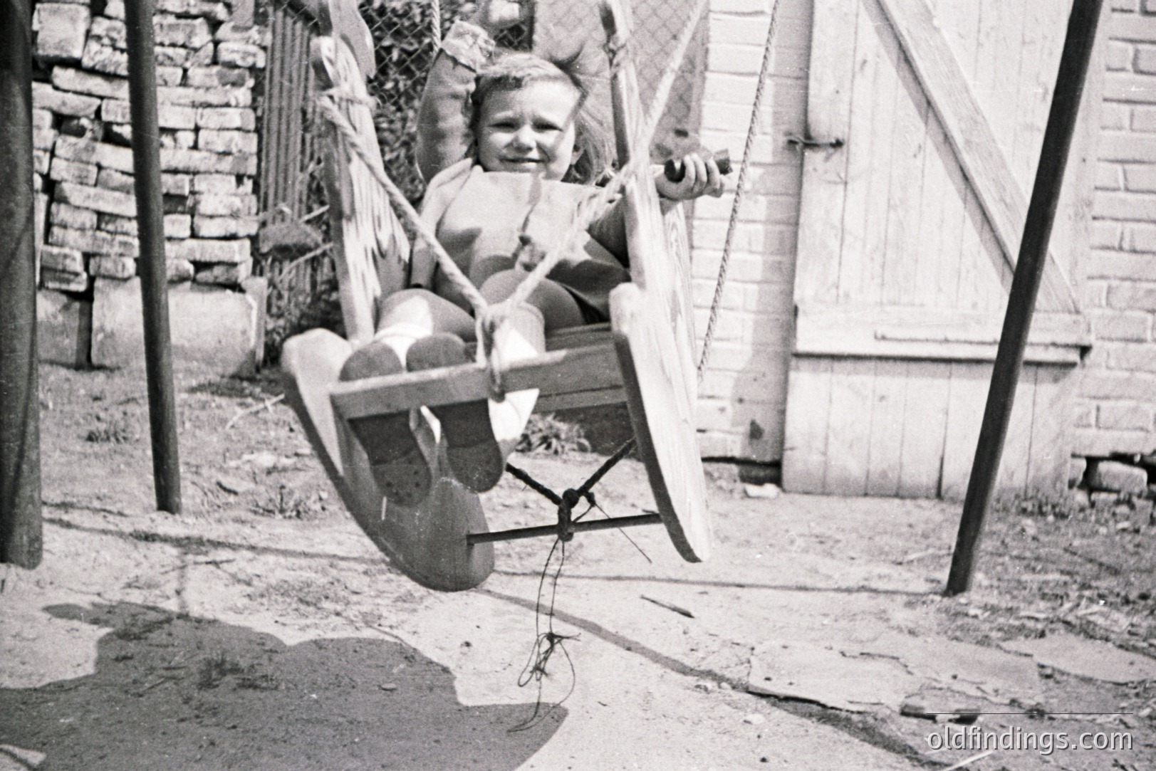 A young child sits on a wooden seat swing, mid-motion, against a backdrop of weathered stone and a clapboard wall. The swing’s construction suggests a handmade or vintage design. Likely 1950s-1960s family snapshot. Possibly an outdoor play area or backyard setting.