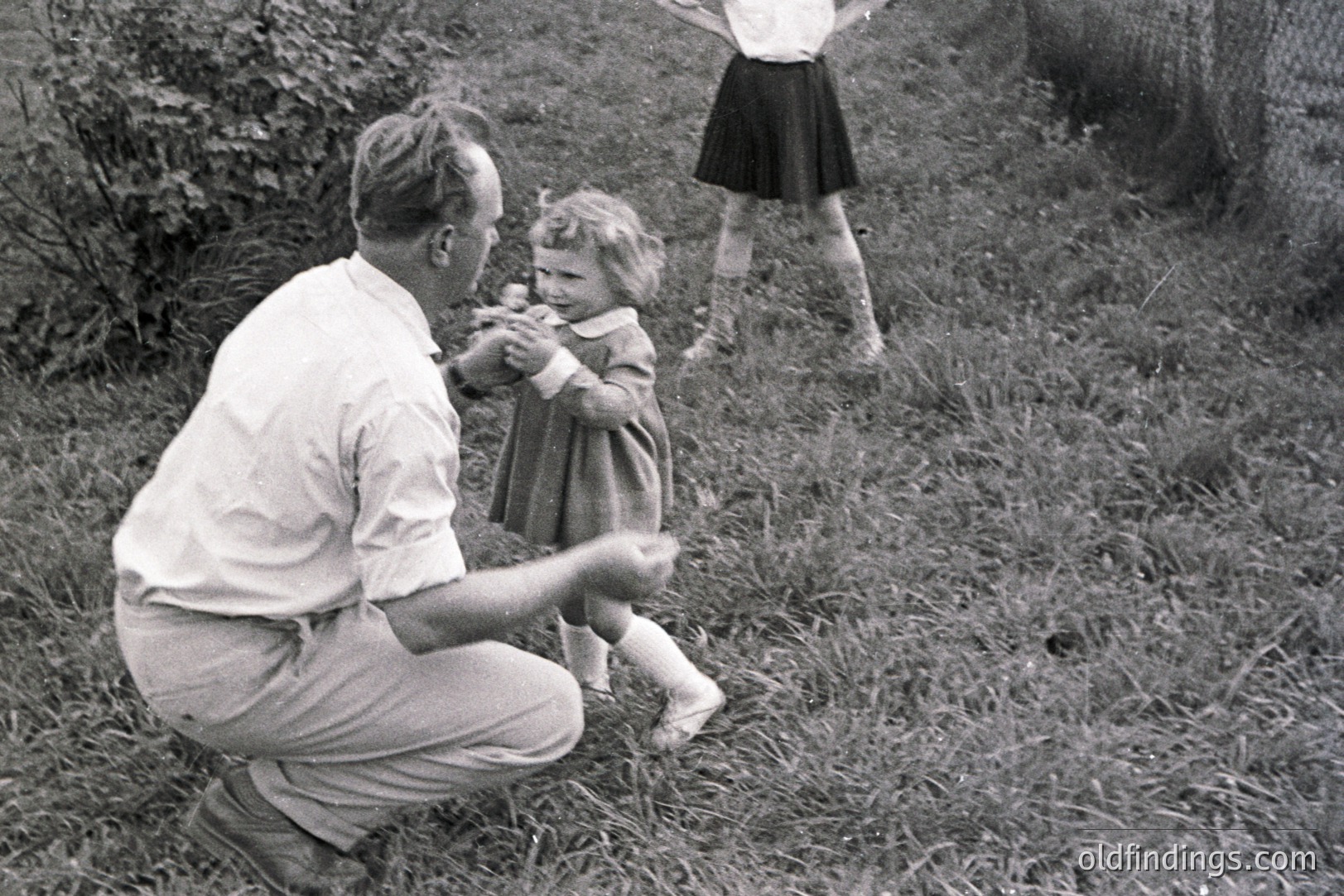 A man in trousers and shirt kneels playfully with a young girl in a dress and socks amidst tall grass. A second child stands in the background. Likely a candid snapshot capturing a family moment in an outdoor setting. Mid-century, possibly 1950s.