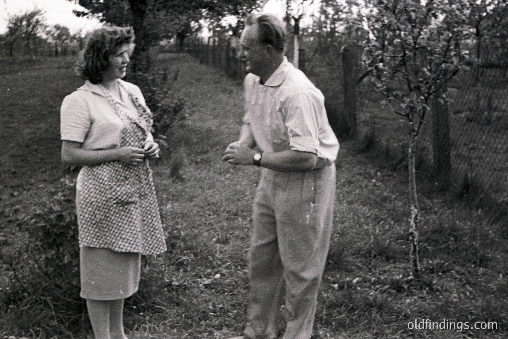 A couple appears to be finishing orchard work. The woman, in a patterned dress & apron, cradles her stomach. The man wears workwear & a wristwatch. Surrounded by blossoming fruit trees. Likely a rural, domestic scene, possibly 1950s. Captures an intimate moment of labor & family.