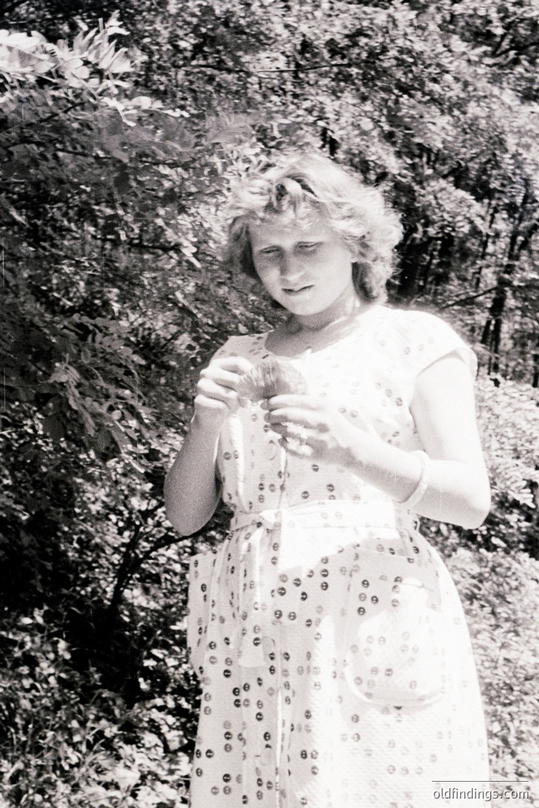 Young woman stands amongst dense foliage, wearing a patterned, short-sleeved dress with a belt. She holds a small, light-colored object, possibly fruit, and appears to be examining it. Likely a casual portrait from the 1960s-1970s, indicative of mid-century fashion.