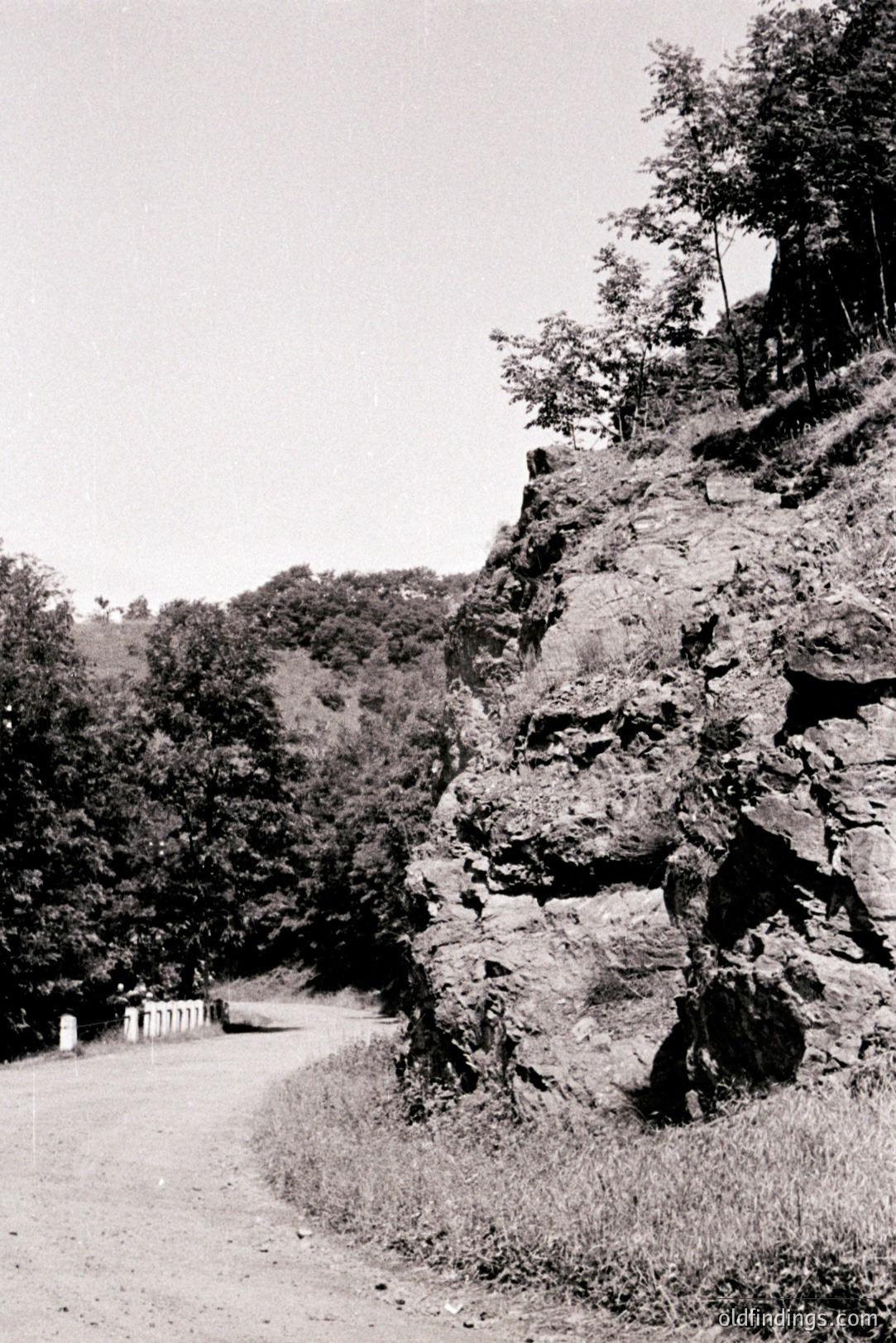 A weathered road curves into a forested area; a rocky cliff face dominates the right foreground. Sparse vegetation grows along the road’s edge. The photo’s style suggests a mid-20th century travelogue or scenic document. Useful for illustrating natural landscapes or early road construction.