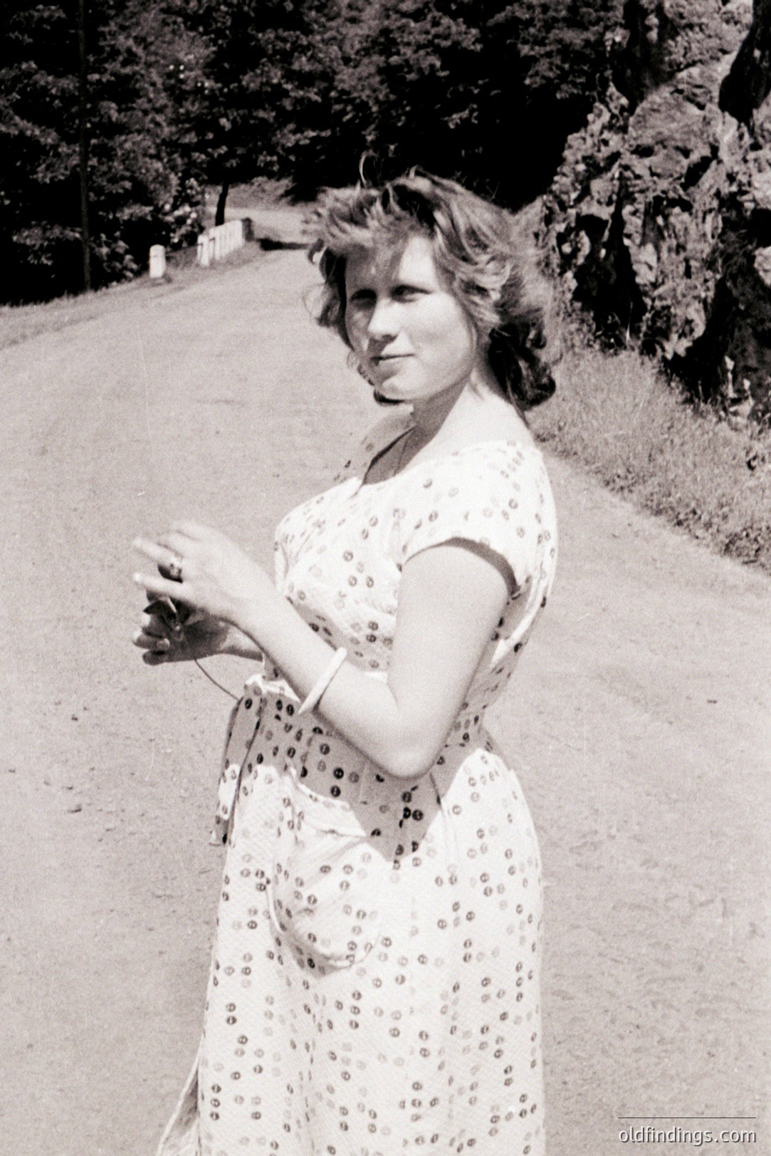 Woman in a floral print dress stands on a paved road, smiling slightly. She wears a bracelet and necklace. Appears to be a candid, mid-century portrait, likely 1960s. Natural lighting. Simple, elegant style. Photographic reference for fashion and portraiture.