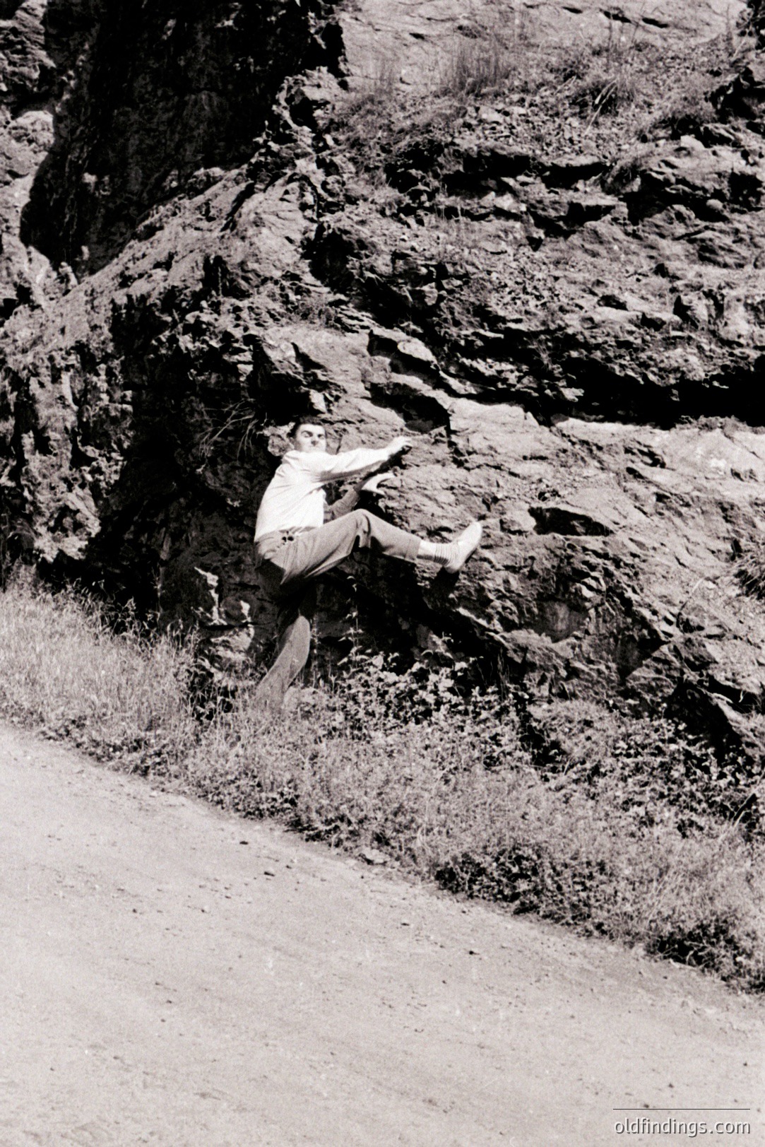 A mid-century man in a suit and dress shoes leaps in a dynamic pose against a rugged, rocky hillside. The rocky terrain with sparse vegetation suggests a natural, outdoor setting, possibly a coastal region. Appears to be a candid, informal portrait.