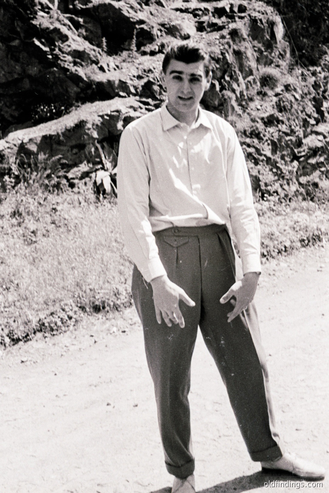 A young man in a crisp, collared shirt and wide-legged trousers stands outdoors, seemingly on a rocky path. He gestures with one hand, displaying a ring. The background features a gentle slope of vegetation and rock formations. Likely 1950s or 60s. A classic, stylish portrait.