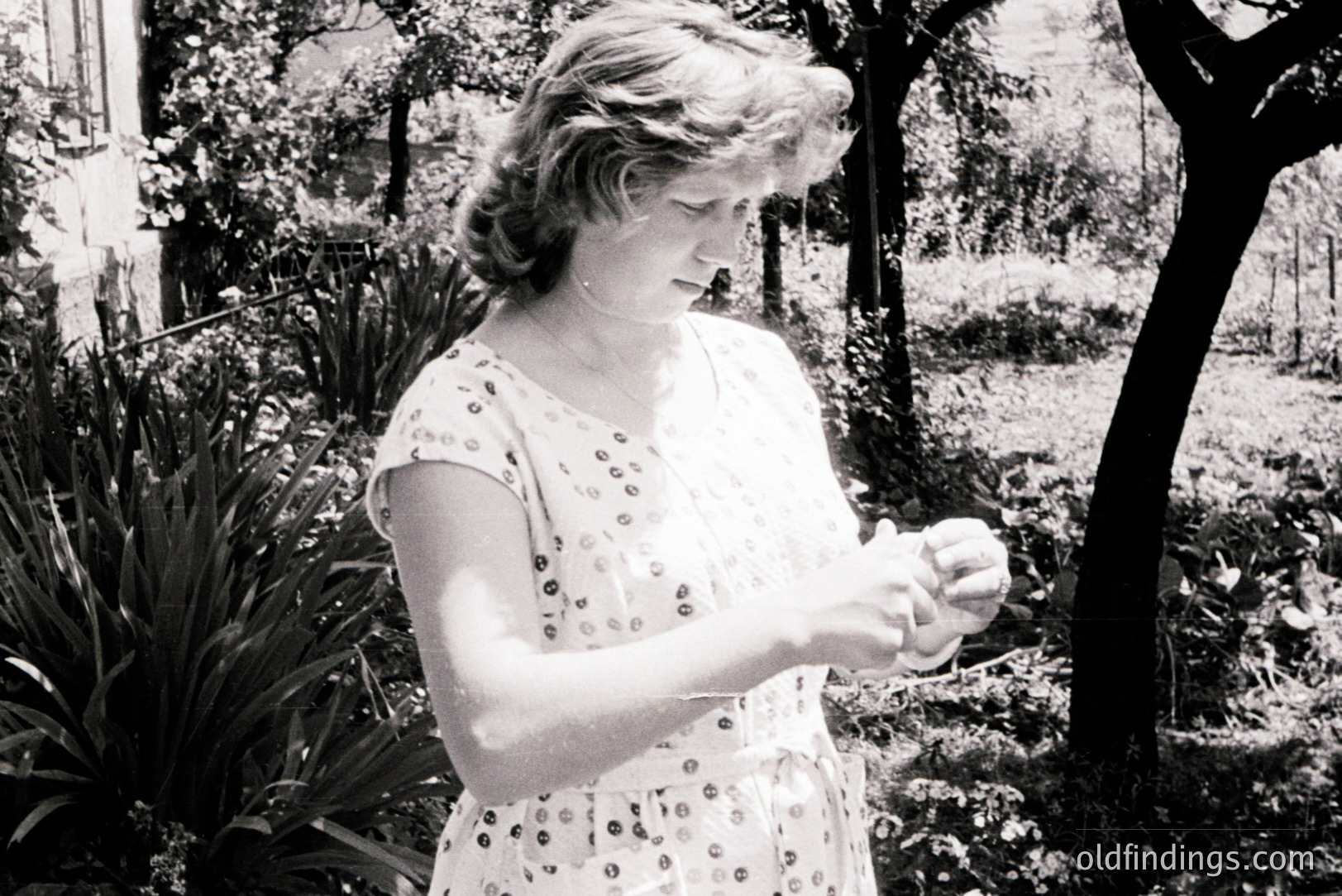 Black and white photo shows a woman in a floral patterned dress, examining an object in her hands. Set in a garden with lush foliage and trees. Likely mid-1970s, based on dress style and photographic quality. Portrait conveys a contemplative mood. Possible stock photo reference.