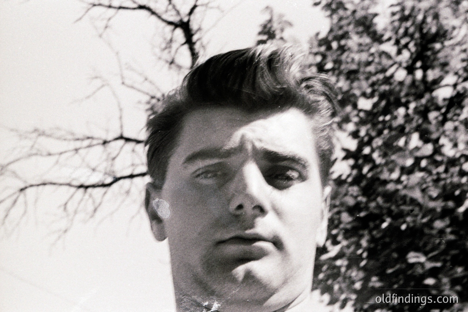 Close-up portrait of a young man with a sharply styled pompadour hairstyle, captured in a low-angle, black-and-white photograph. Minimalist background suggests outdoor setting. Likely a mid-20th century, possibly 1950s style. Good for retro design or historical portraiture.