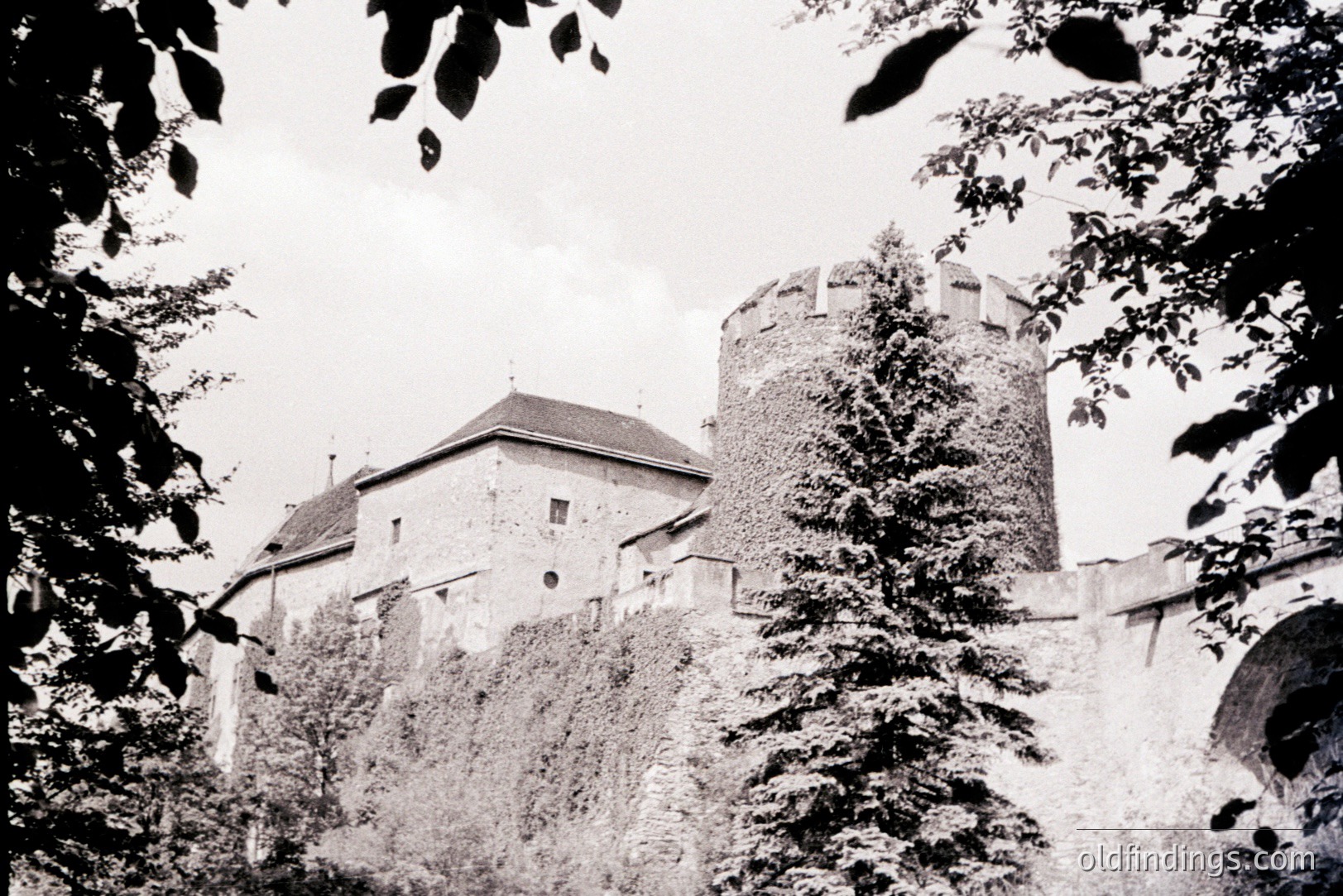 A stone castle with a rounded tower, partially covered in ivy, rises above a stone wall. Architecture suggests a medieval or Renaissance European style. Likely a defensive structure, potentially from the 16th-18th centuries. High commercial value for historical research/stock imagery.