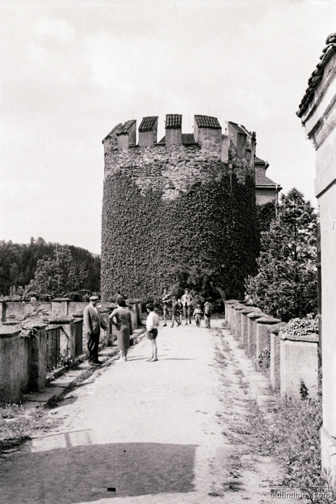 A walled courtyard path leads towards a weathered, ivy-covered stone tower. Several figures, including children, are visible along the path. The tower’s crenellations suggest medieval origins. Likely taken in the mid-20th century, possibly Eastern Europe. A detailed architectural reference.