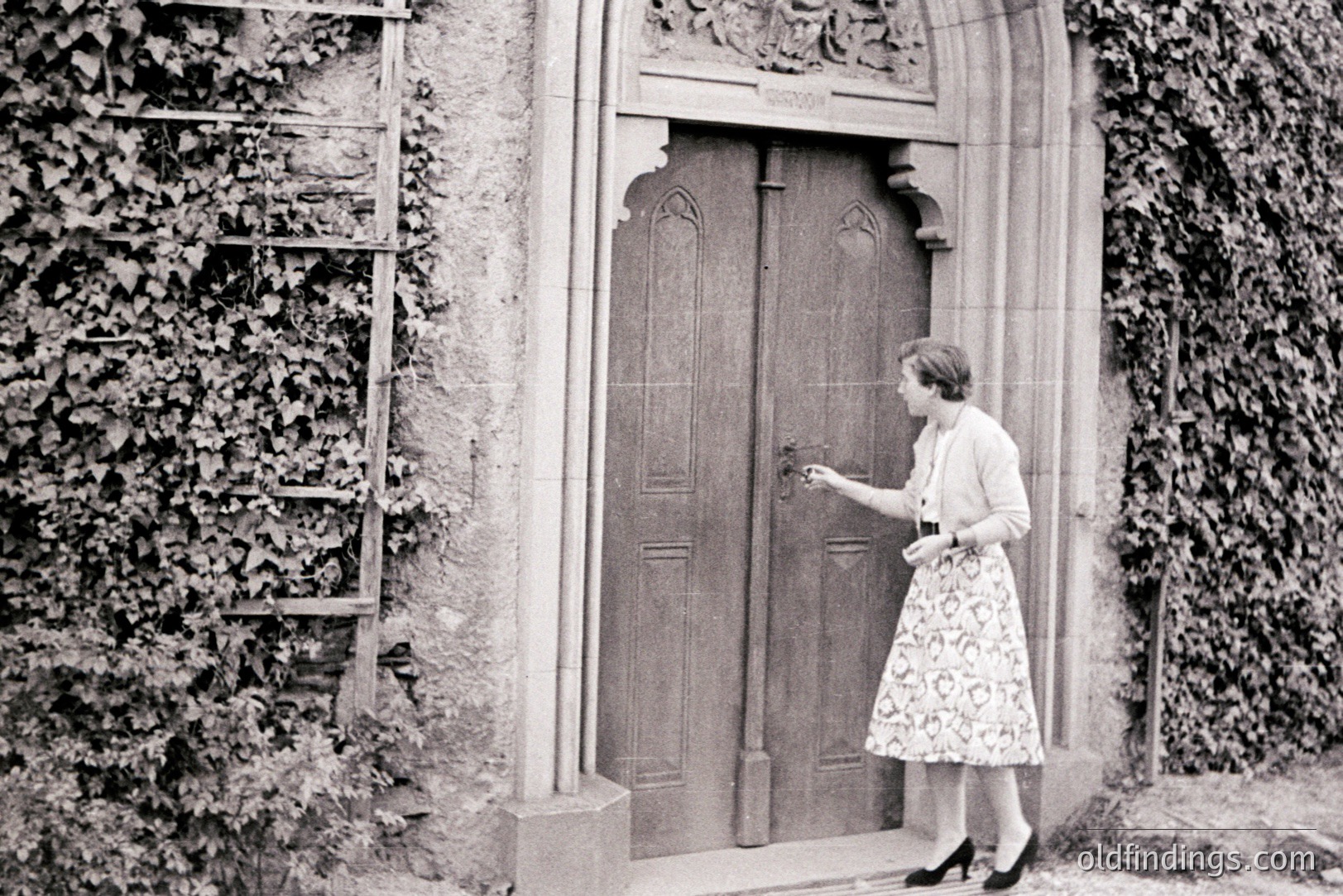 A woman in a patterned skirt suit reaches for a wrought-iron door handle within a stone archway, ivy-covered wall to her left. Architectural details suggest a European location, possibly a manor house or historic building. Style and clothing indicate the 1950s or 1960s. Likely a candid, documentary-style image.
