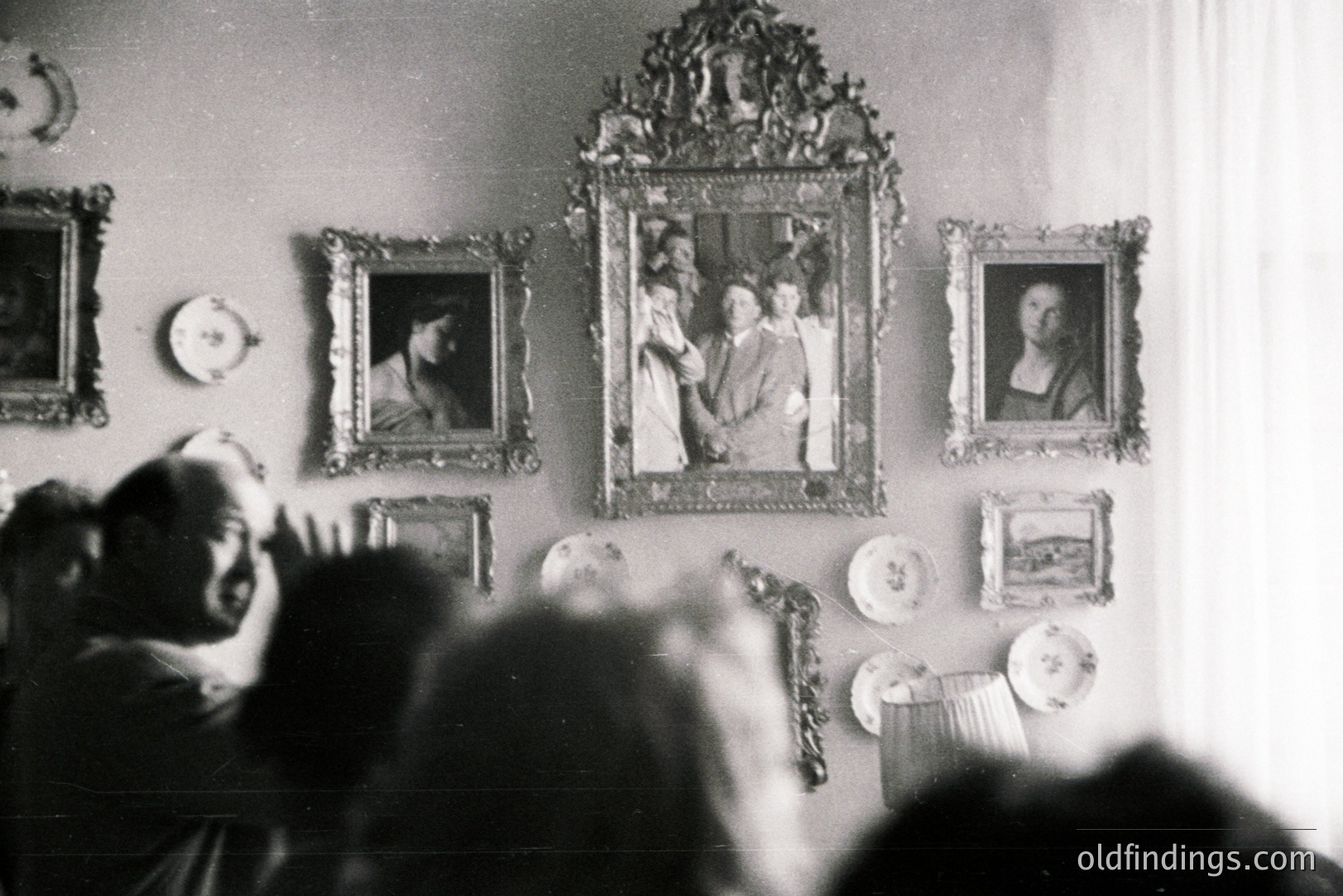 Framed portraits & decorative plates adorn a wall, visible through a blurred foreground of onlookers. Formal portraiture, ornate gilt frames, and delicate porcelain suggest a well-appointed interior, possibly a stately home. Likely mid-20th century. Could serve as design reference.