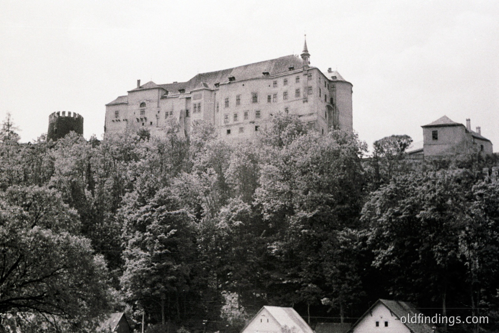 Grand Baroque chateau rises above lush foliage. A fortified tower is visible on the left. Likely 18th century, European architectural style. The structure exhibits multiple windows and a central spire. Possible estate or castle grounds.