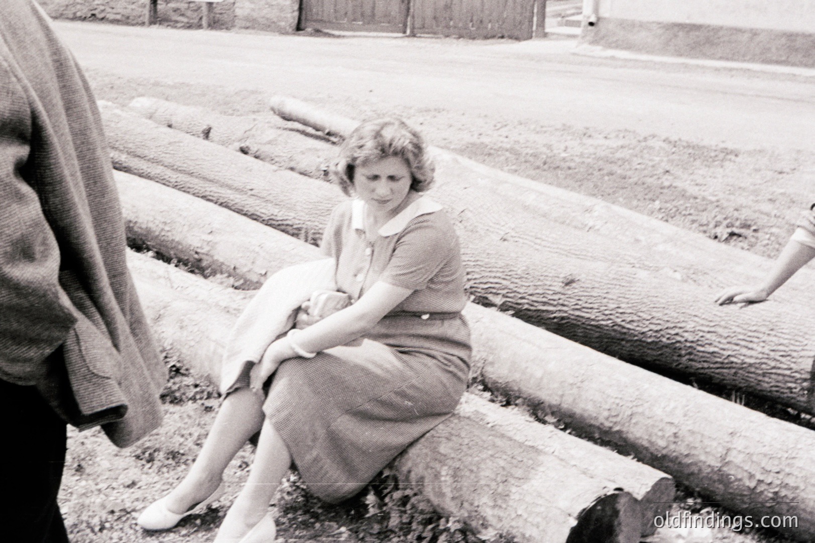 A woman in a 1960s style dress and heels sits on stacked logs beside a street. Two other figures are partially visible. The scene suggests rural or industrial location, possibly post-war rebuilding. Architectural details hint at Eastern European style. Documentary-style photograph.