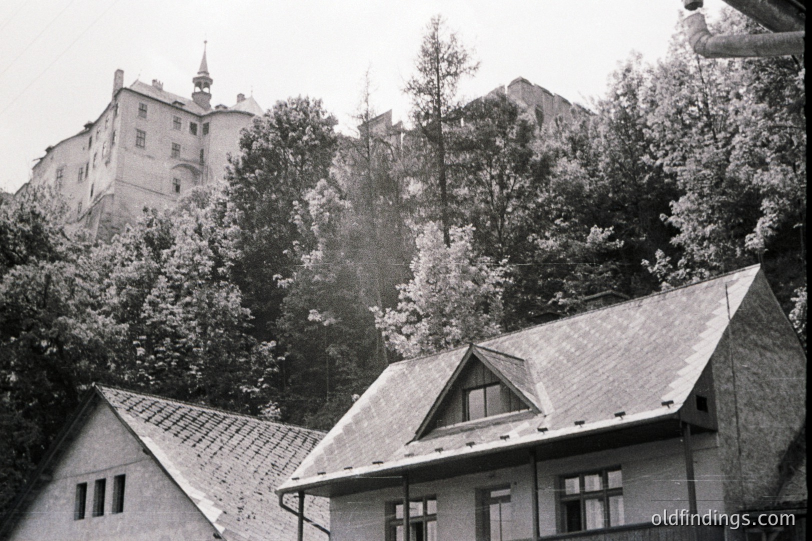 Roofed homes frame a view of a medieval castle perched on a wooded hillside. Classic gabled architecture with tile roofs and small windows suggest a European village. The image’s grainy quality hints at a mid-20th century snapshot. Possibly Eastern Europe.