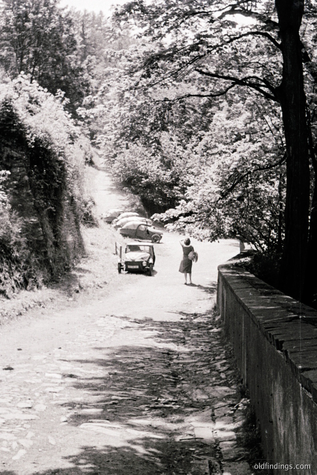 Winding, narrow road flanked by a concrete retaining wall and dense trees. Three vintage automobiles are visible, with a woman in a coat walking away from the viewer. Likely a rural European location, mid-20th century. Possible stock photo reference for period travel or automotive advertising.