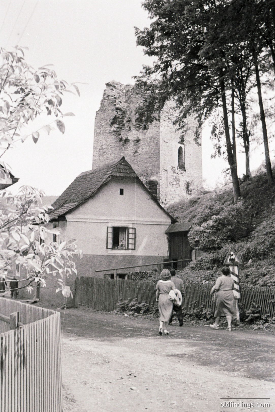 Rustic village scene: three figures stroll along a dirt road past a traditional, tiled-roof house adjacent to the ruins of a stone tower. Appears to be a rural landscape with dense foliage. Likely central Europe, 1950s-1970s. Potential for design references.
