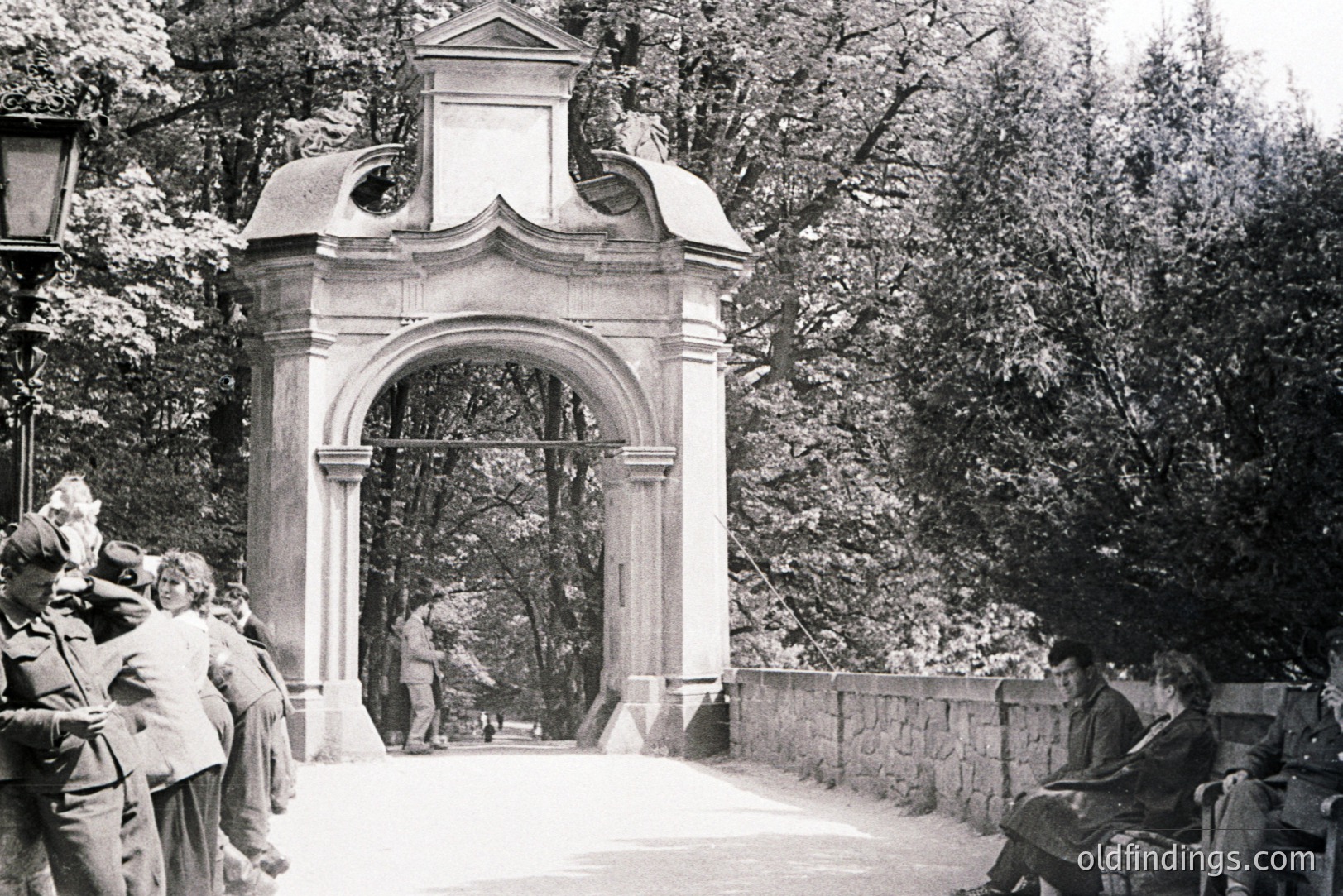 Monochrome image of a monumental stone archway with figures on a pathway & stone retaining wall. Several formally dressed individuals populate the foreground and distant background. Likely a park or garden setting. Architectural detail suggests Central or Eastern European origin, c. 1910-1930. Good potential for design/heritage projects.